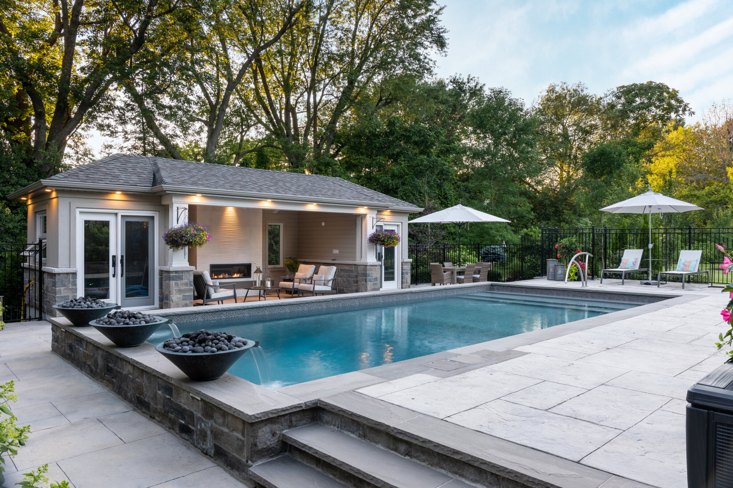 Luxurious backyard with a swimming pool, patio area, sun loungers, umbrellas, a pool house with a fireplace, trees for privacy, and decorative bowls on pedestals.