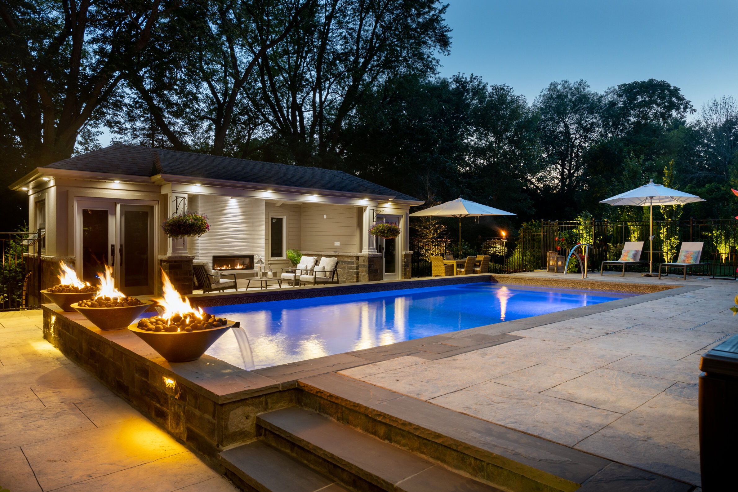 Luxurious backyard at dusk, featuring a lit swimming pool, fire bowls, patio furniture, and ambient lighting, with no people visible.