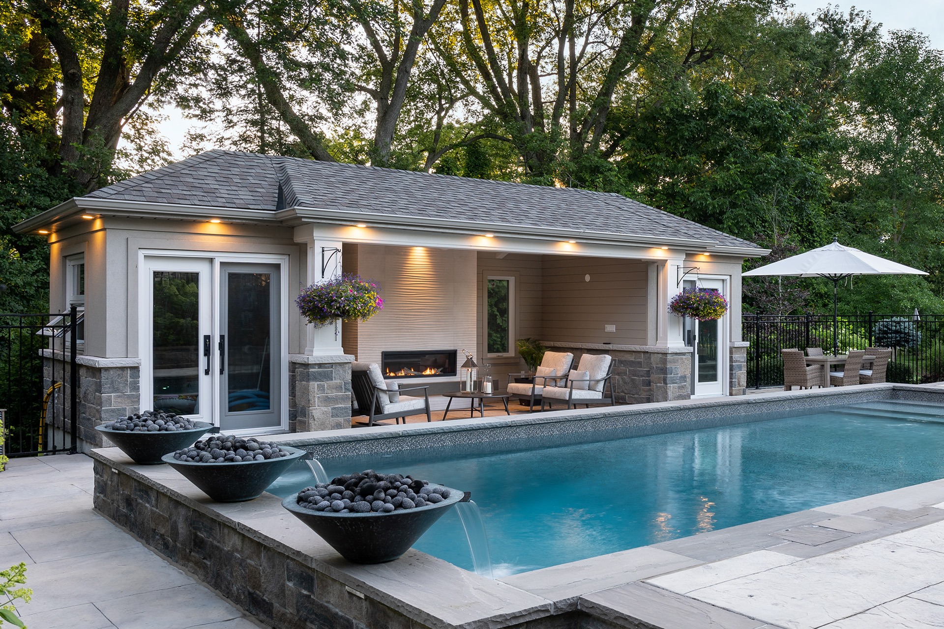 An elegant poolside setting with a cozy pavilion, outdoor furniture, a fireplace, and water features, surrounded by trees at dusk.