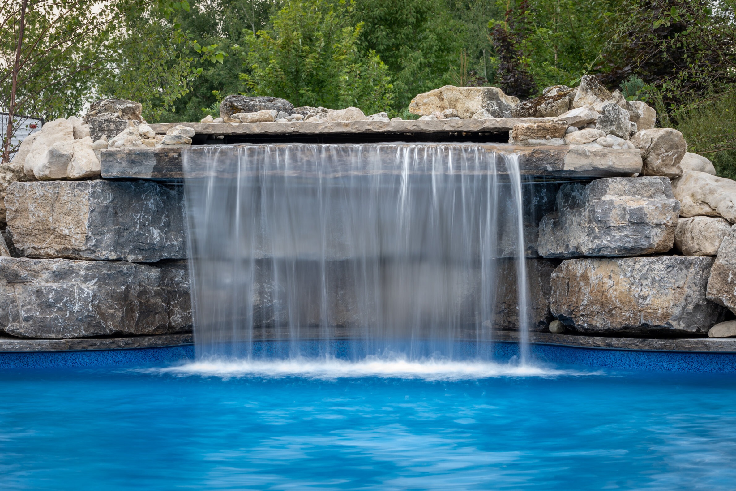 A serene artificial waterfall cascades over textured rocks into a bright blue pool, photographed with a slow shutter speed for a smooth water effect.