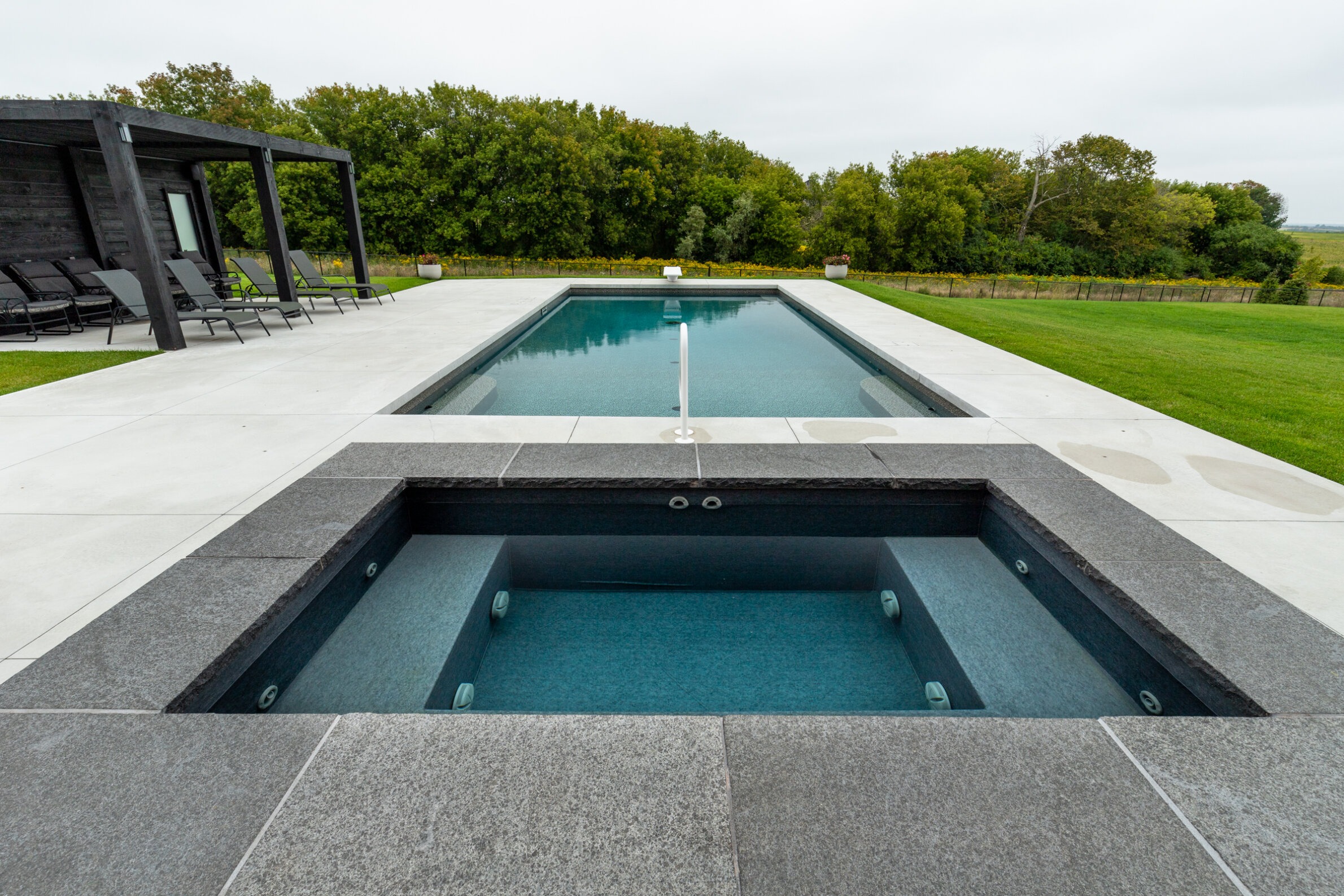This image shows a modern outdoor pool and a hot tub with grey stone decking, lounging chairs to the side, and greenery in the background.