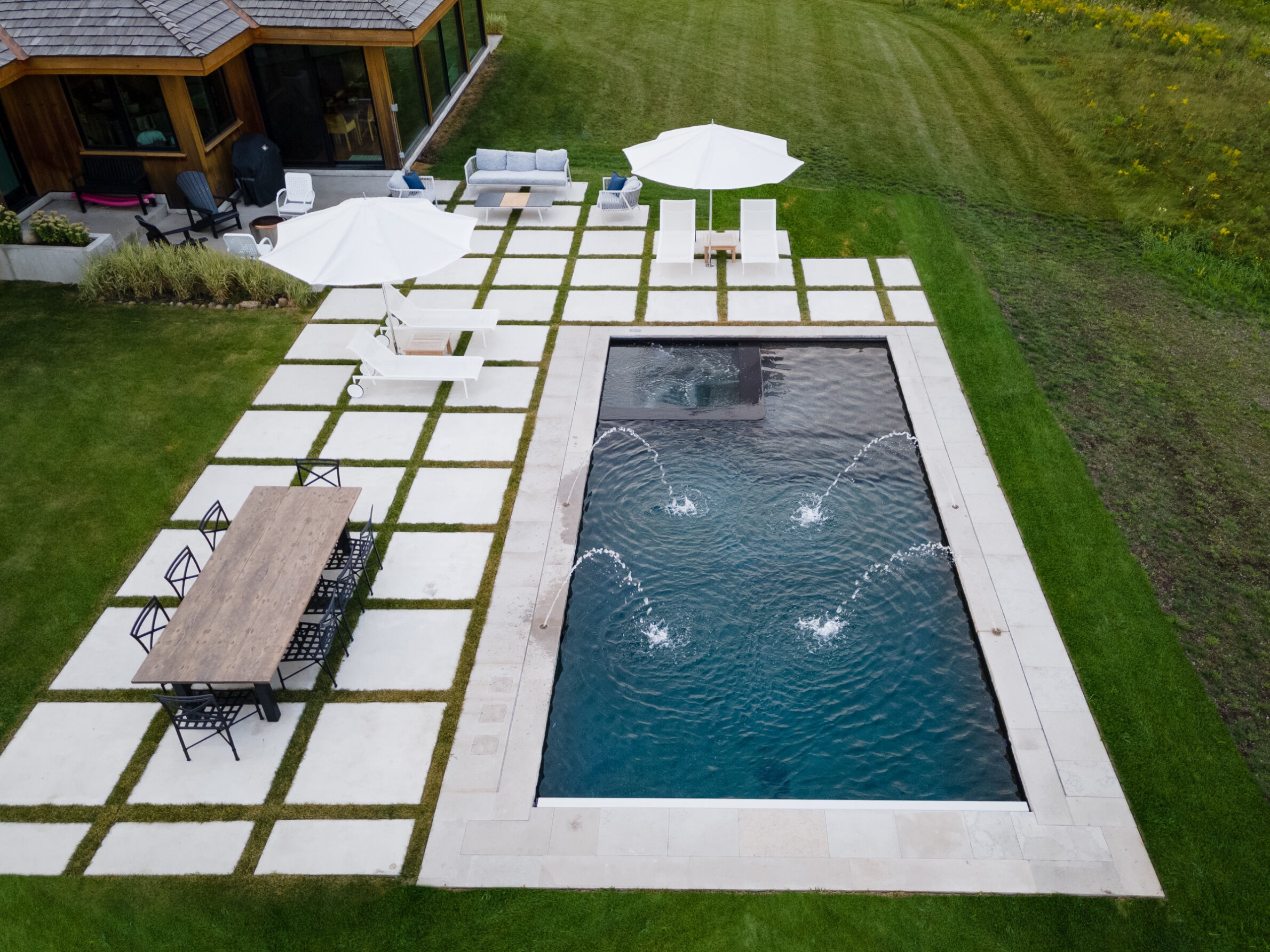 Aerial view of a backyard with a rectangular pool, fountain jets, a wooden dining table, sun loungers, umbrellas, and manicured grass with a house adjacent.