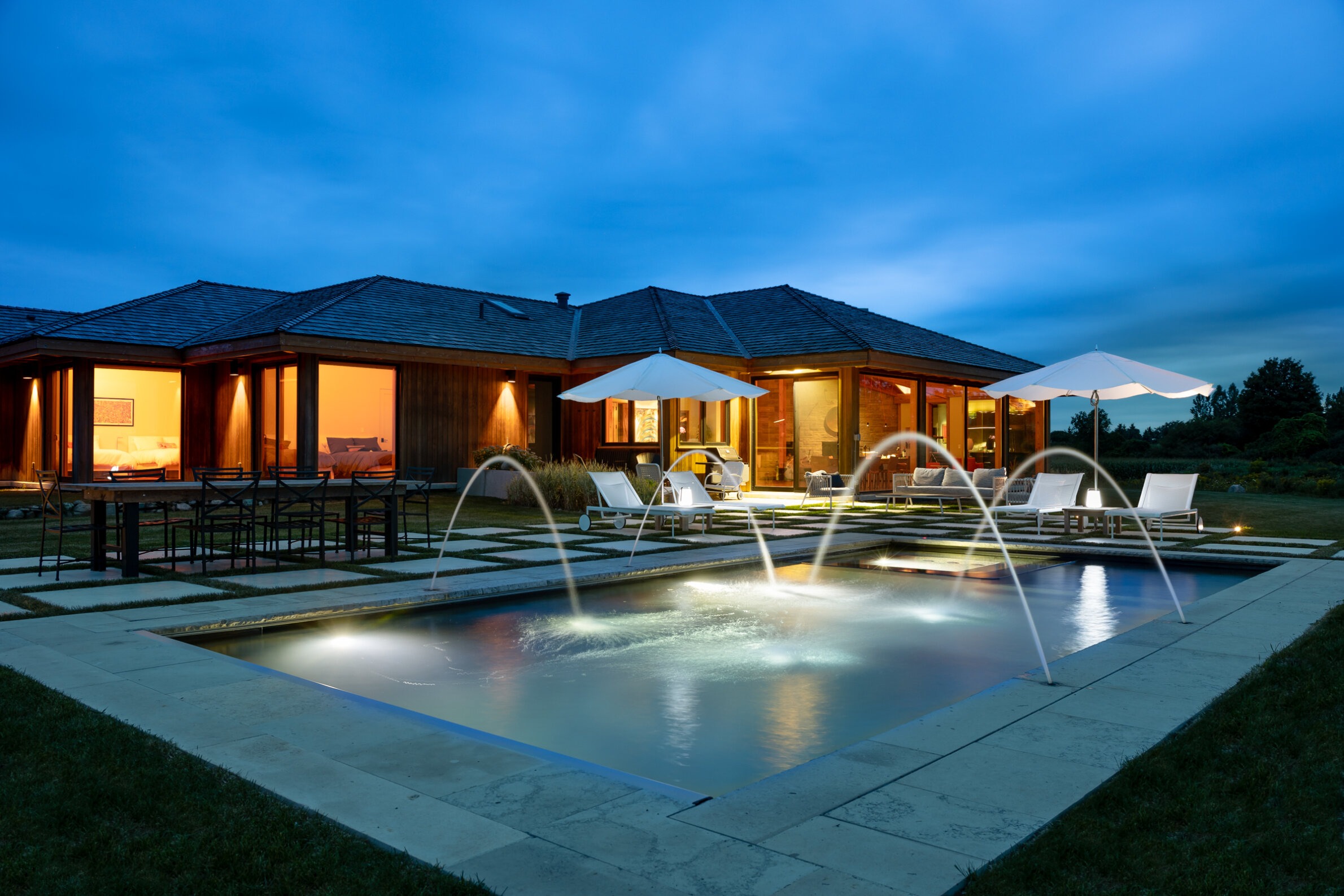 A modern house with illuminated interiors, a swimming pool with fountain jets, lounge chairs, and outdoor lighting during twilight. Tranquil and luxurious setting.