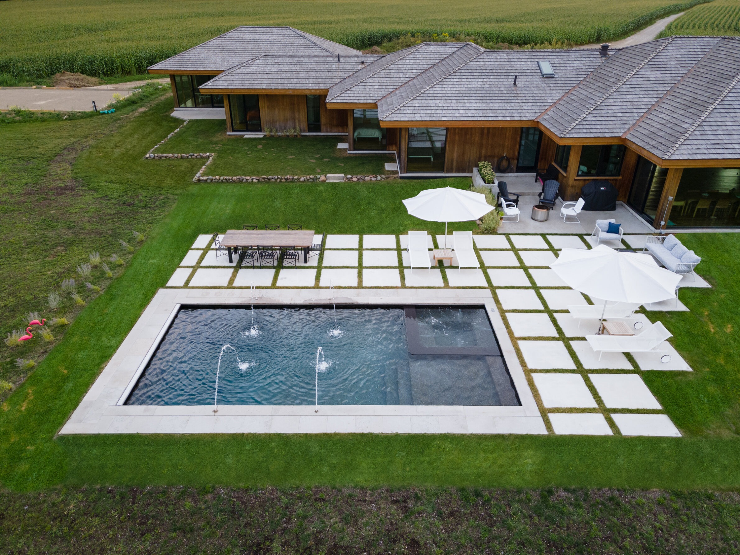 Aerial view of a modern house with a rectangular pool, outdoor seating, umbrellas, surrounded by green lawns and adjacent farmland.