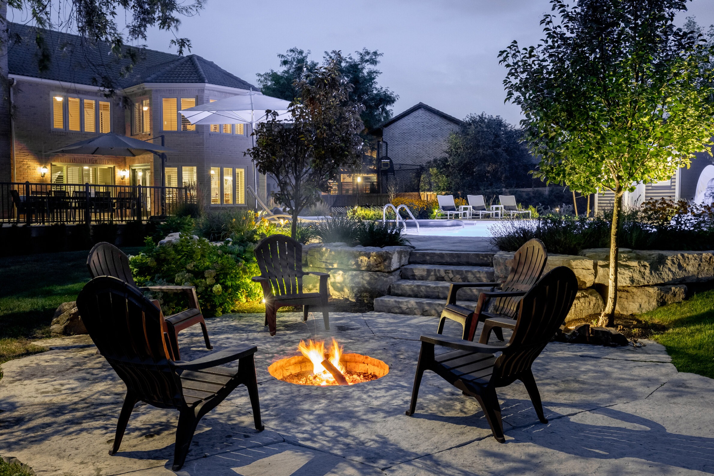 An evening backyard setting with a lit fire pit, Adirondack chairs, a pool, a two-story house with illuminated windows, and surrounding greenery.