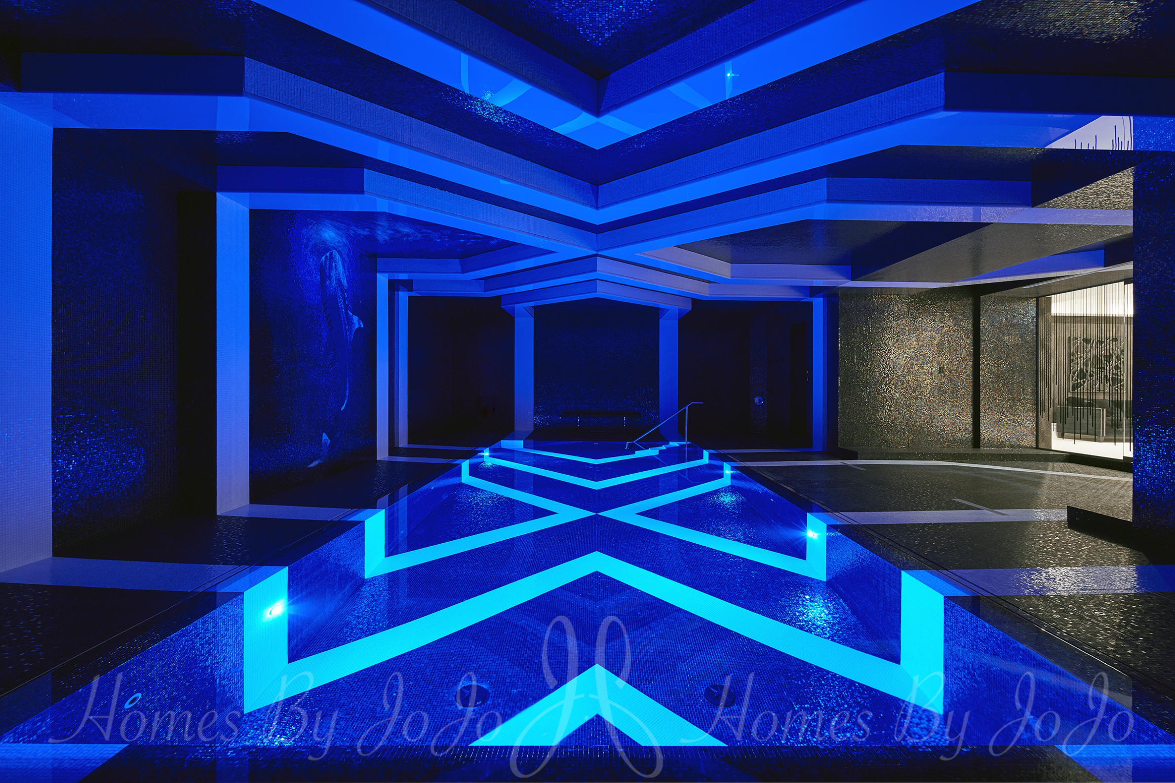 A night time view of an indoor swimming pool with glowing, bold geometric patterns surrounded by raised, light colored geometric lines on dark walls