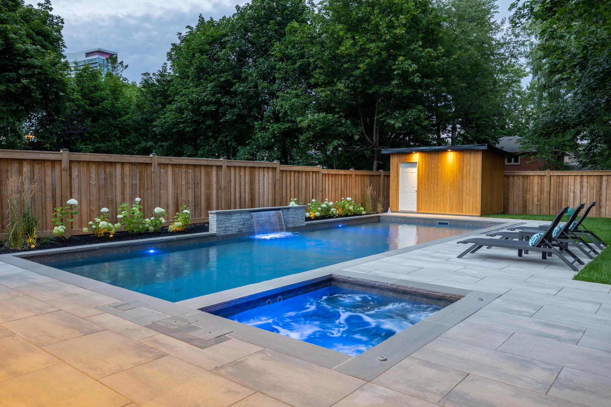 A serene backyard with a swimming pool and hot tub at dusk. Stone patio, wooden fence, greenery, and a small building with a lit interior.