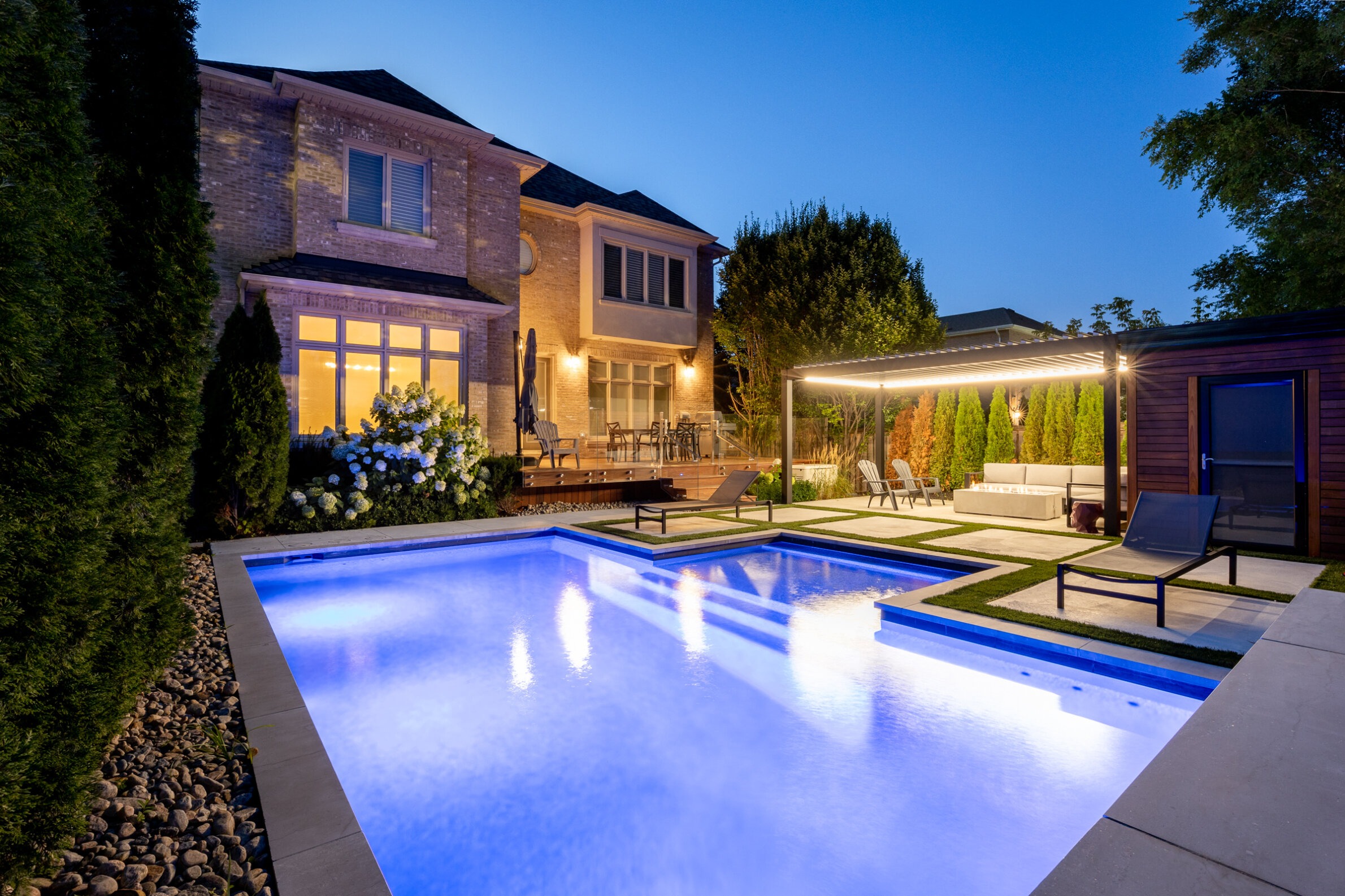 A luxurious backyard at twilight with an illuminated blue swimming pool, elegant furniture, landscaping, and a modern house with warm interior lights.