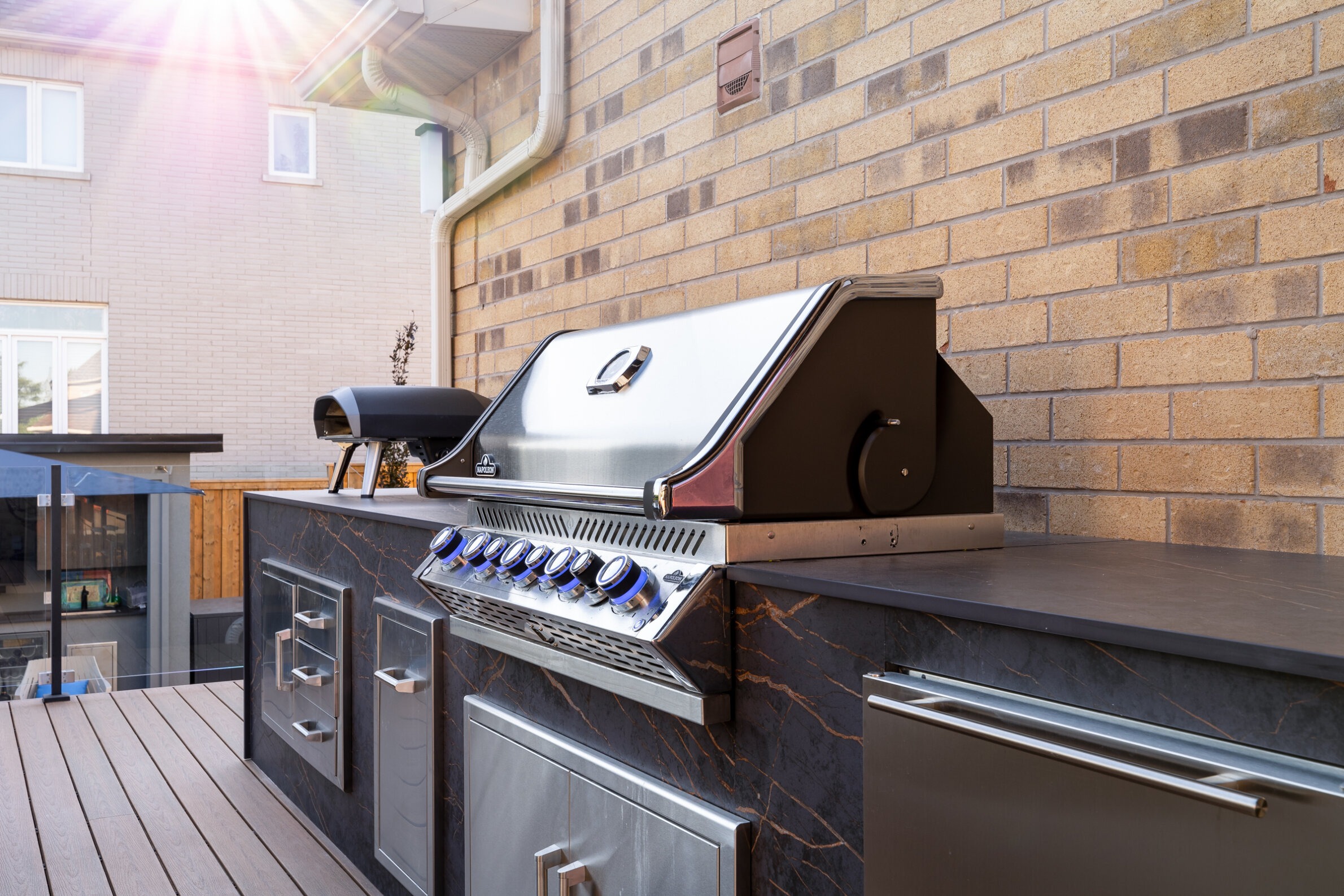 An outdoor kitchen with a modern gas grill and storage cabinets set against a brick house. Sunny weather casts a warm glow.