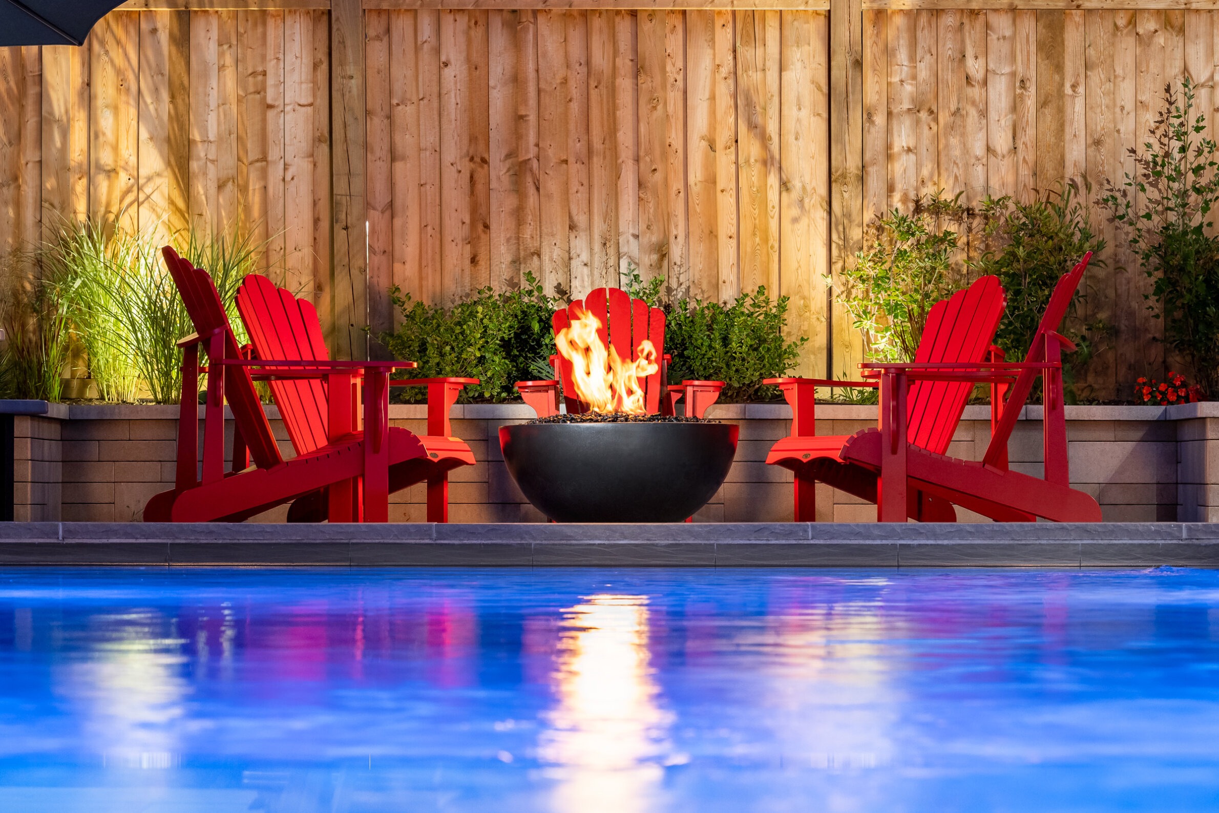 Two red Adirondack chairs face a fire pit with glowing flames, beside a pool with blue reflections, and a wooden fence background at dusk.