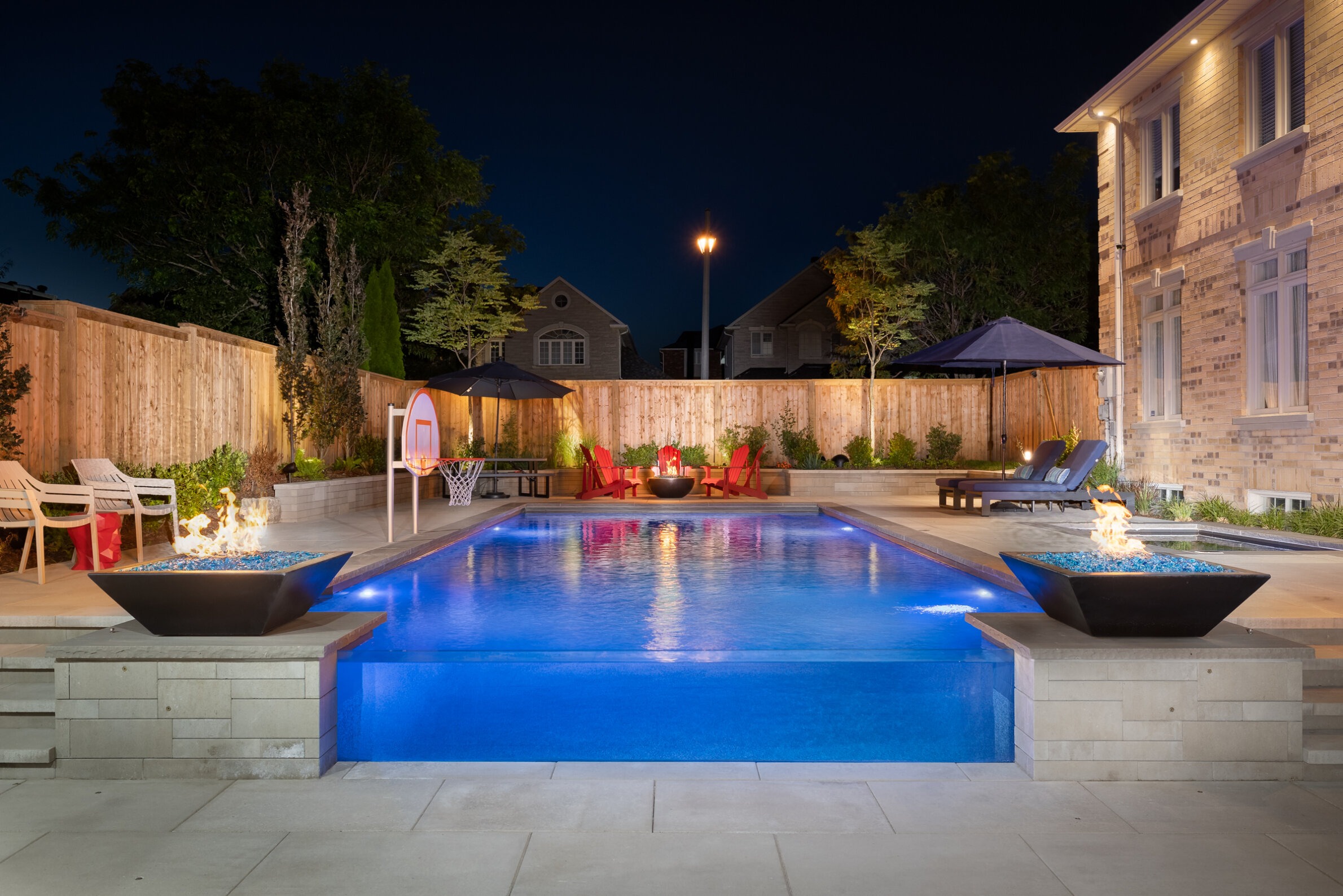 A luxurious backyard with a lit swimming pool at dusk, fire bowls, lounge chairs, basketball hoop, and ambient lighting, enclosed by a wooden fence.