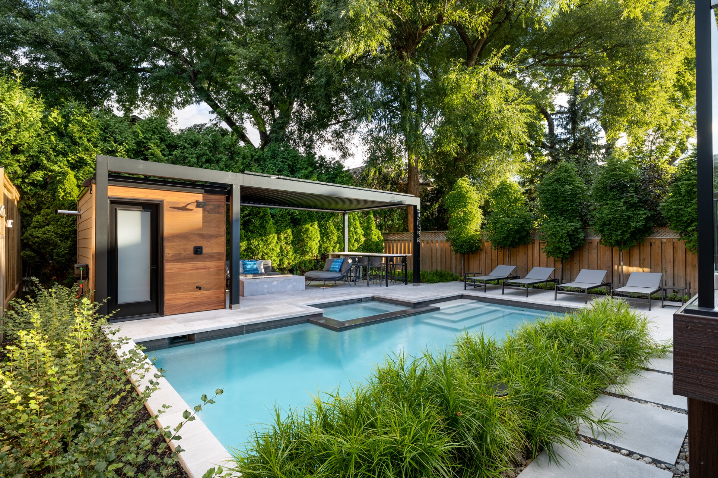 An inviting backyard with a rectangular swimming pool, modern pool house, loungers, green landscaping, and a pergola-covered seating area. Surrounded by a wooden fence.