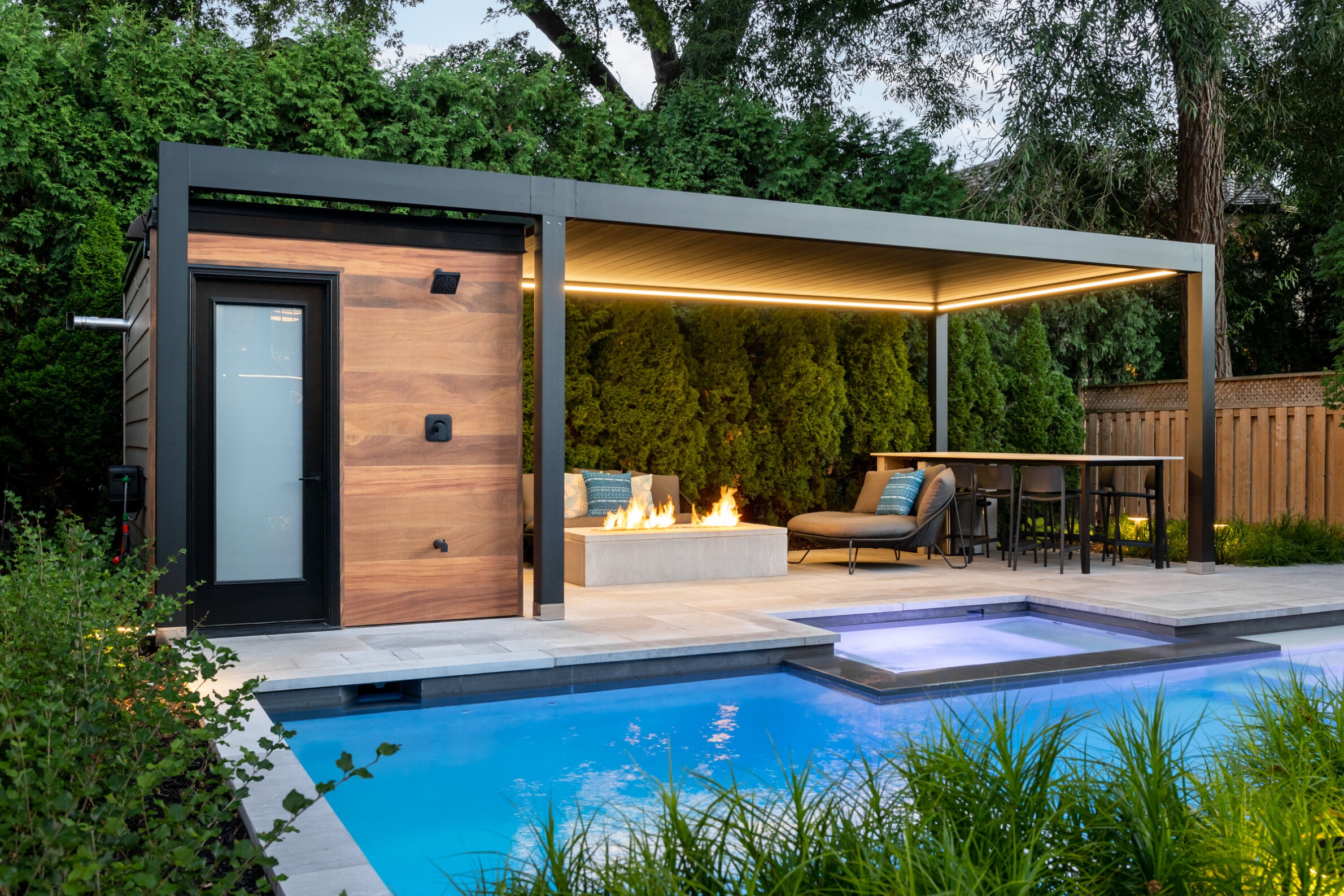 A modern backyard with a swimming pool, covered patio area with a fireplace, lounging furniture, dining set, and lush greenery during the evening.