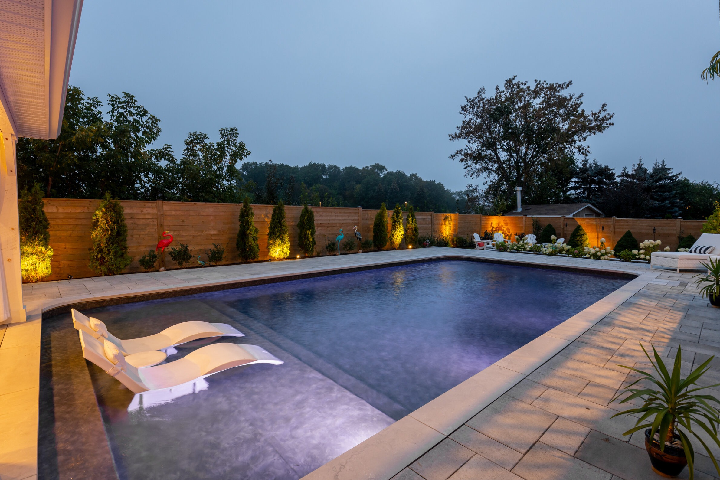 An outdoor swimming pool with evening lighting, surrounded by a privacy fence and landscaping, reflects the sky at twilight. A floating lounger sits on the water.