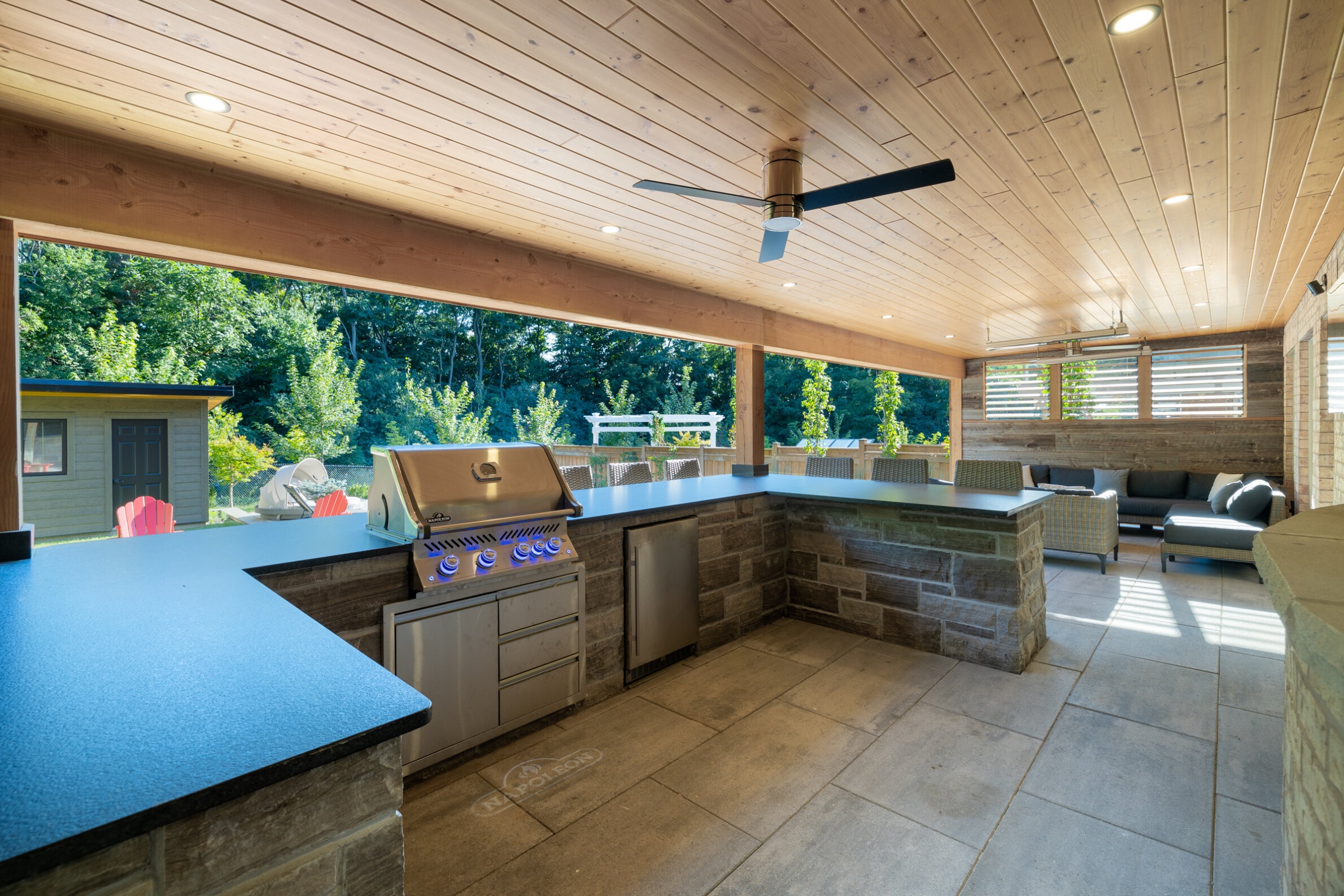 This image shows a spacious, covered outdoor kitchen area with modern appliances, a seating lounge, a wooden ceiling, and a view of a garden.