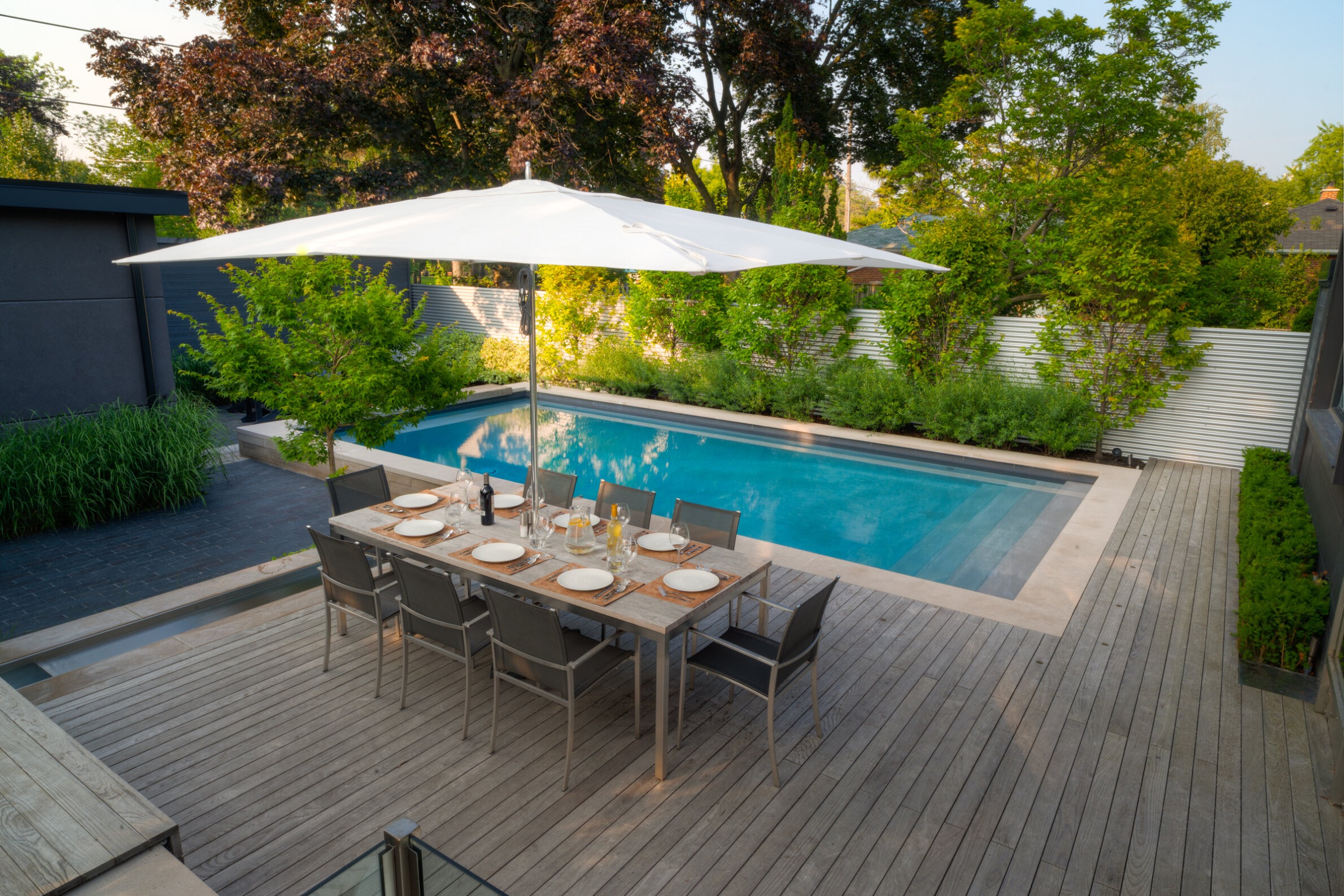 A serene outdoor setting with a dining table set for a meal under a white umbrella, wooden decking, and a rectangular swimming pool surrounded by greenery.