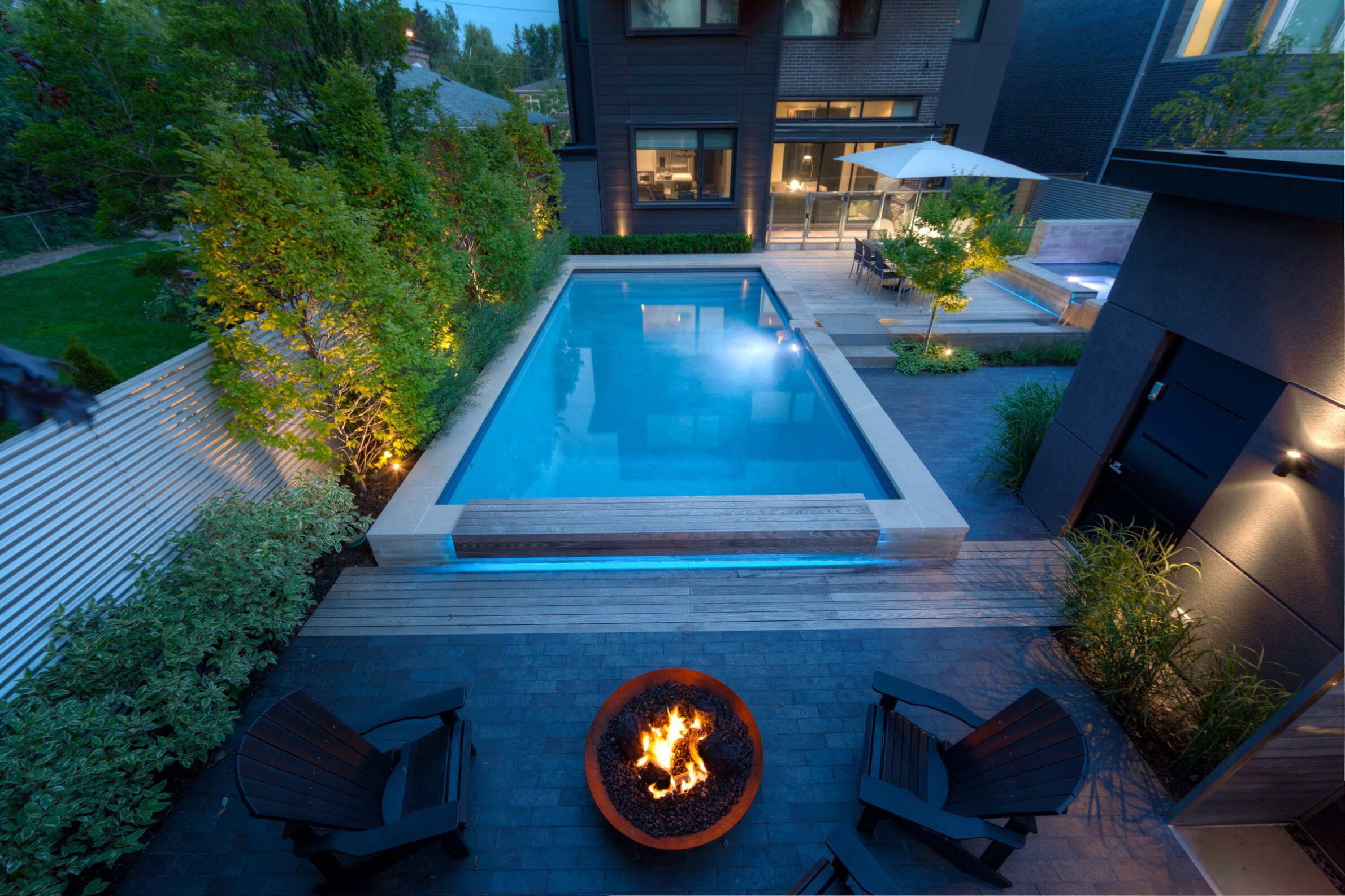 An evening view of a modern backyard with a lit swimming pool, outdoor furniture, a fire pit, and a well-lit patio adjacent to a house.