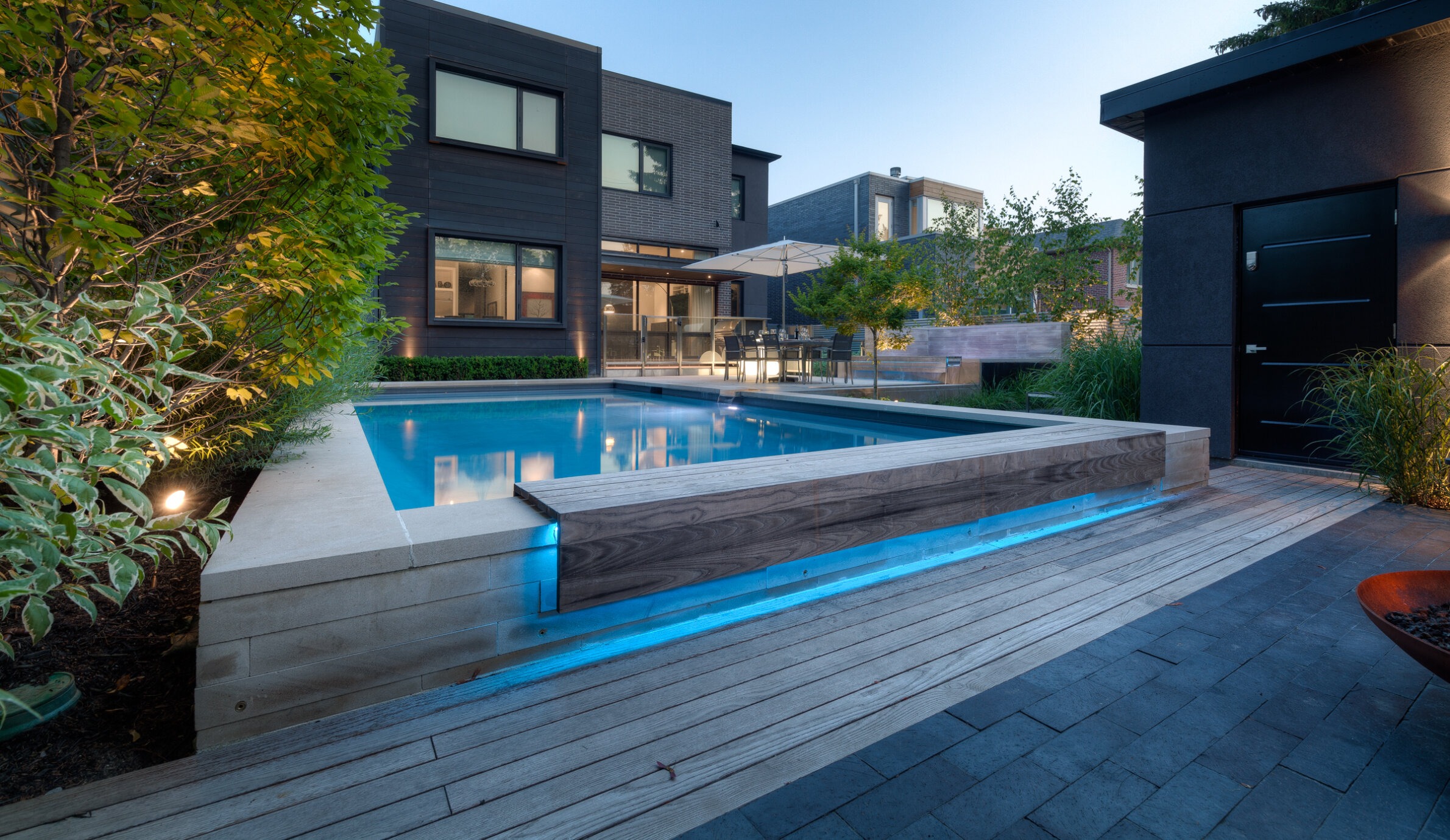 Modern backyard with a rectangular swimming pool featuring underwater lighting. Wooden decking, landscaped garden, contemporary house with outdoor furniture visible in twilight.