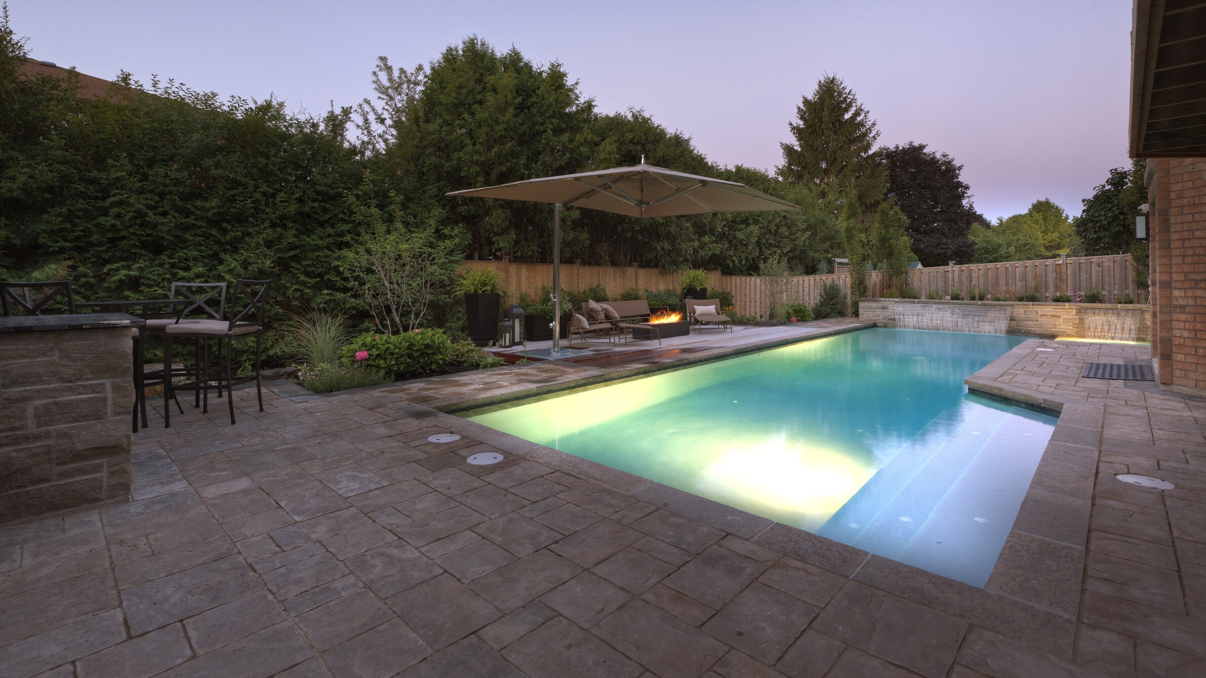An elegant backyard at dusk, featuring a lit swimming pool, outdoor firepit seating area, patio umbrella, landscaping, and a stone patio.
