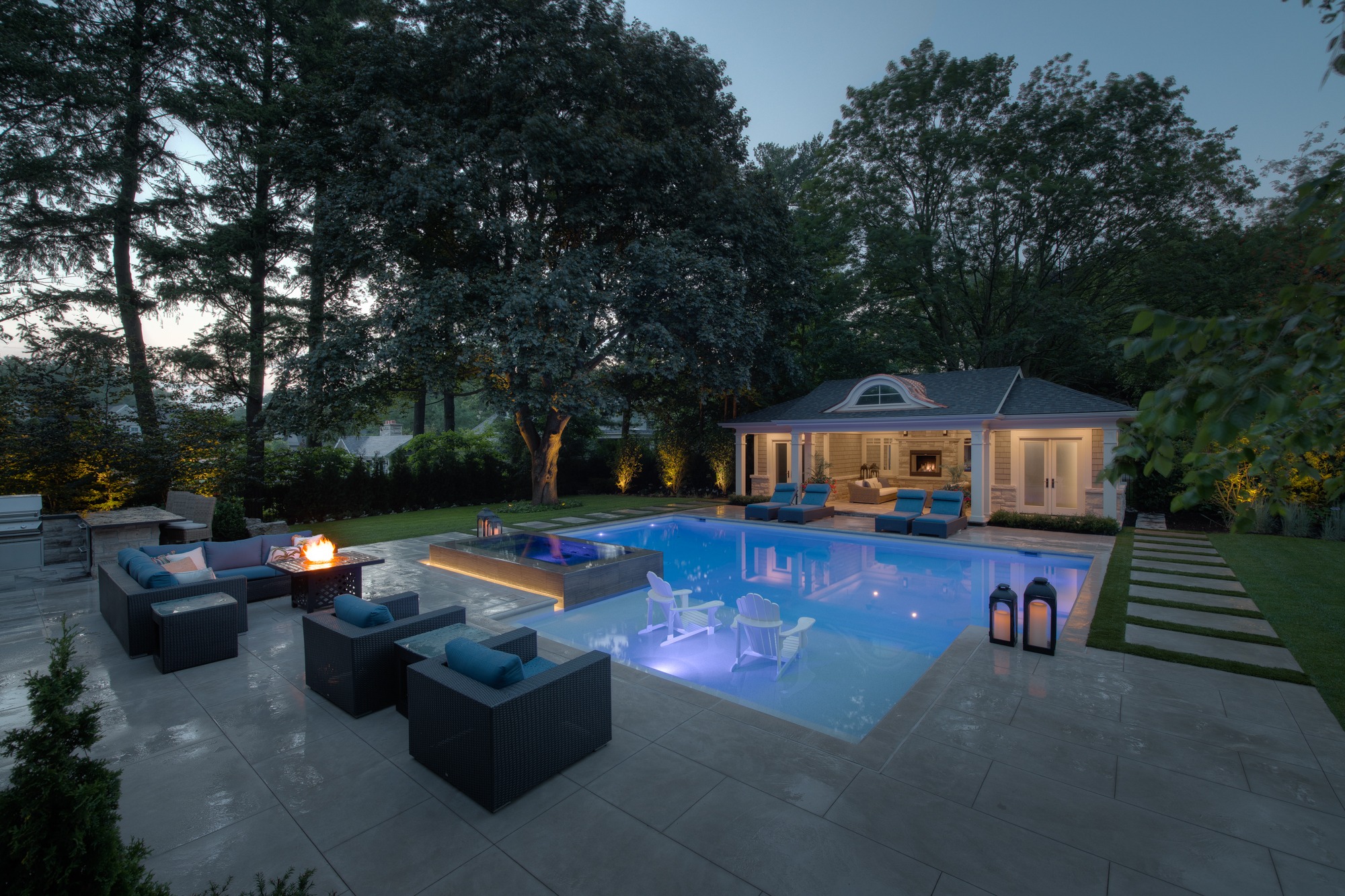 Luxurious backyard with a lit swimming pool at dusk. Outdoor seating, fire pit, and a pool house surrounded by trees provide a tranquil setting.