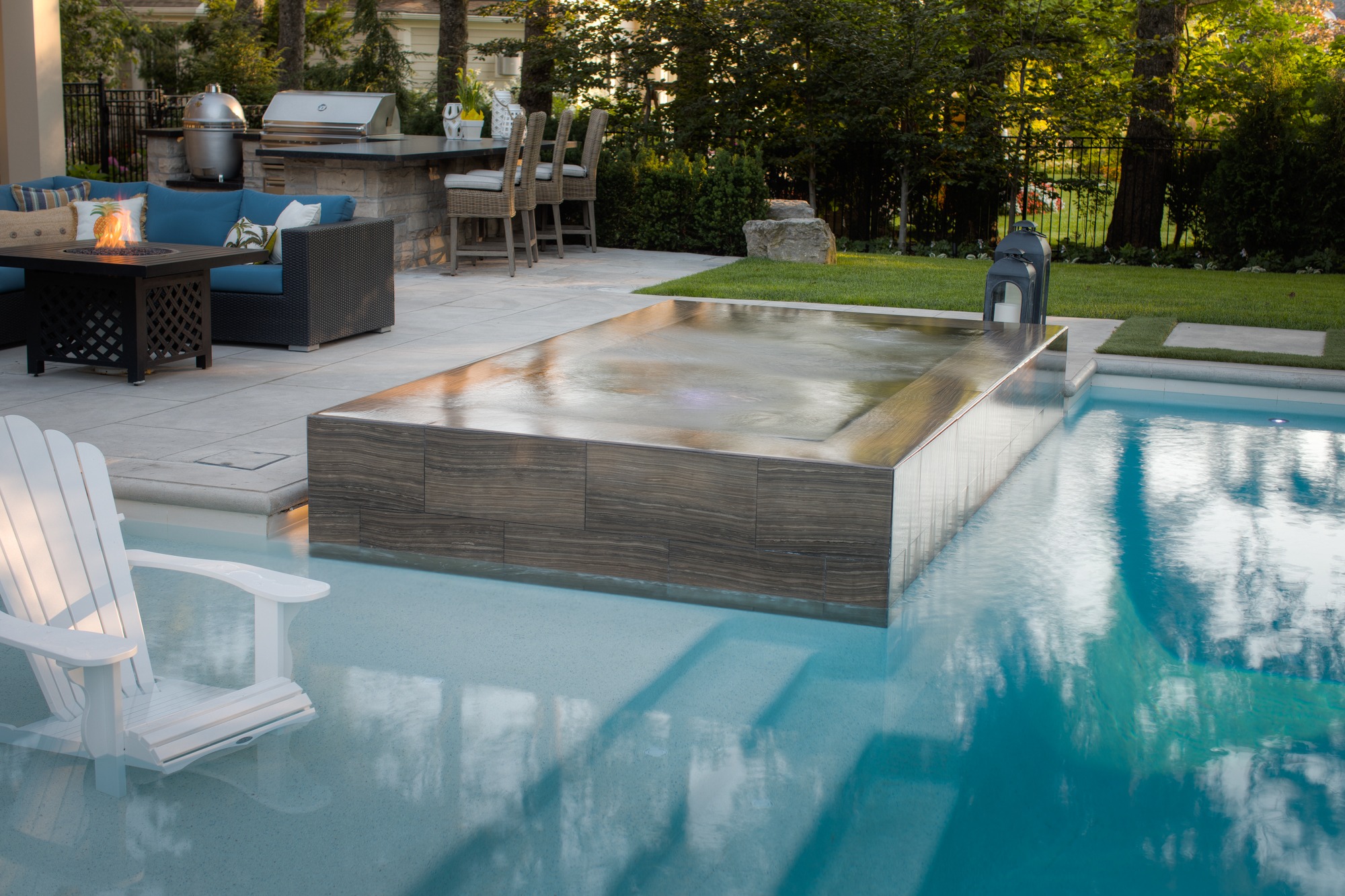 An outdoor residential setting featuring a pool with an integrated hot tub, surrounded by a patio with seating, a fire pit, and a grill area.