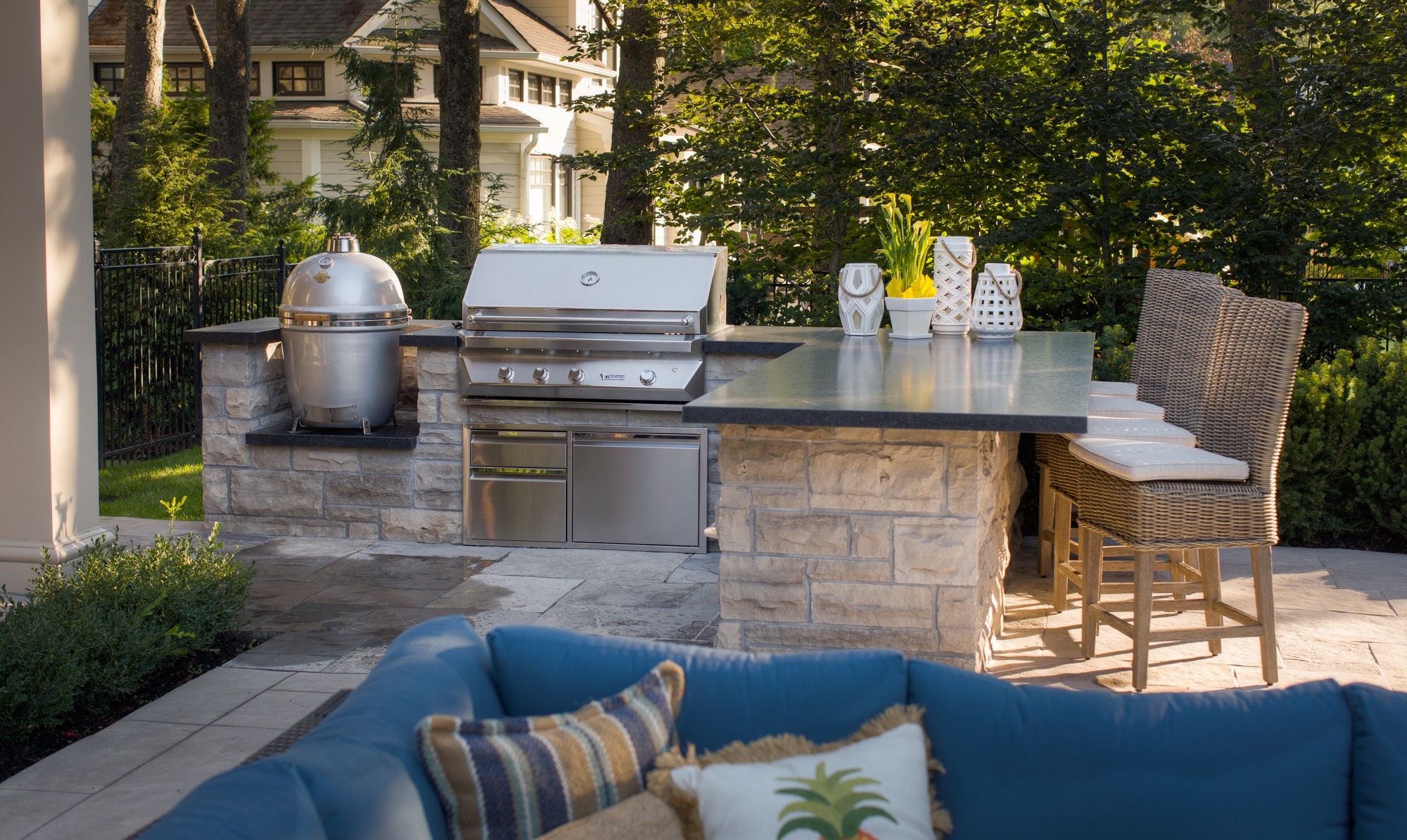 An outdoor kitchen setup with a grill, stone countertop, wicker chairs, decorative items, surrounded by greenery, and part of a seating area.