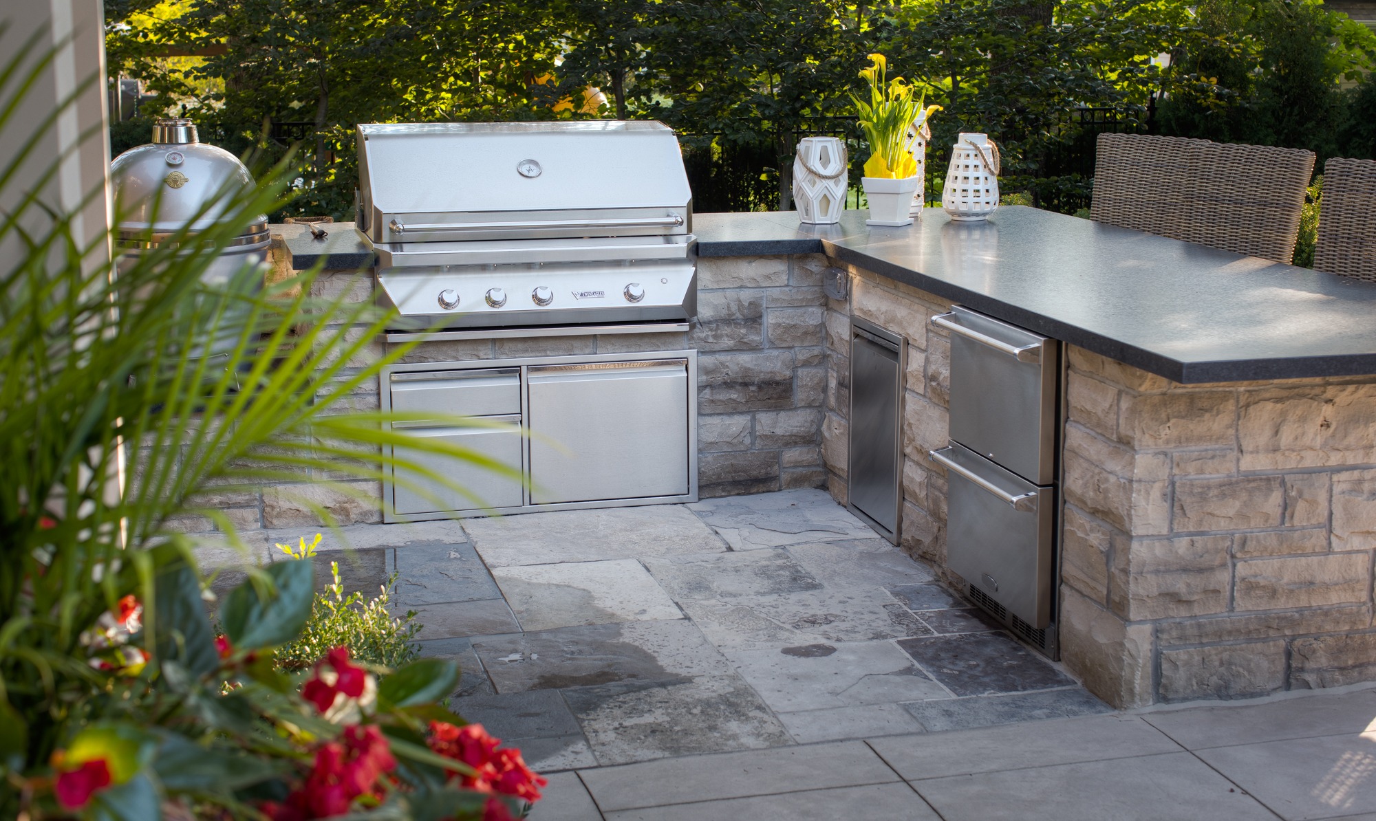An outdoor kitchen with a built-in grill and refrigerator, set in stone counters, surrounded by greenery, flowers in the foreground, on a sunny day.