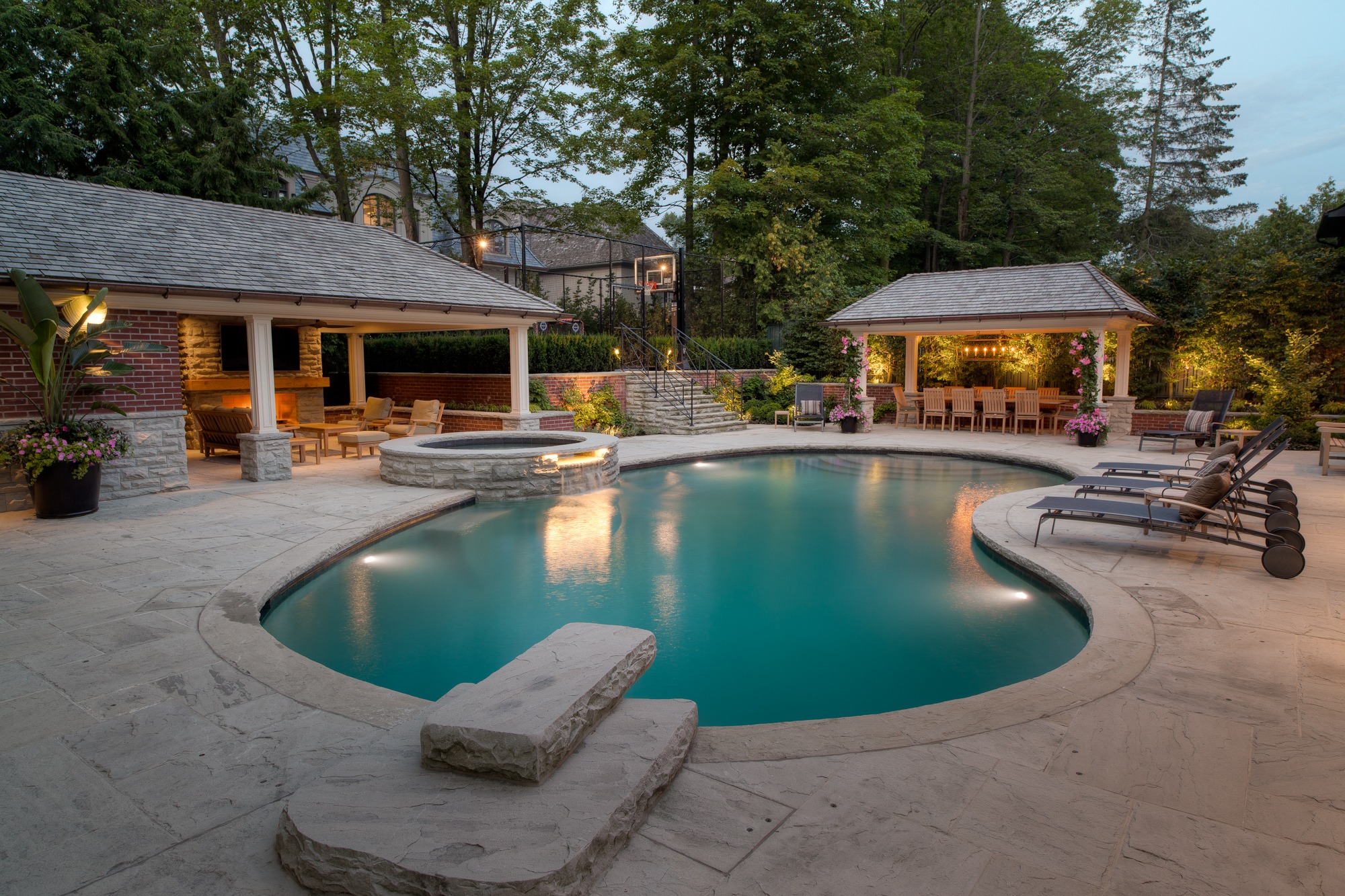 An elegant backyard with a curved swimming pool, surrounded by stone patios, loungers, and covered areas with lighting, trees, and a warm ambiance.