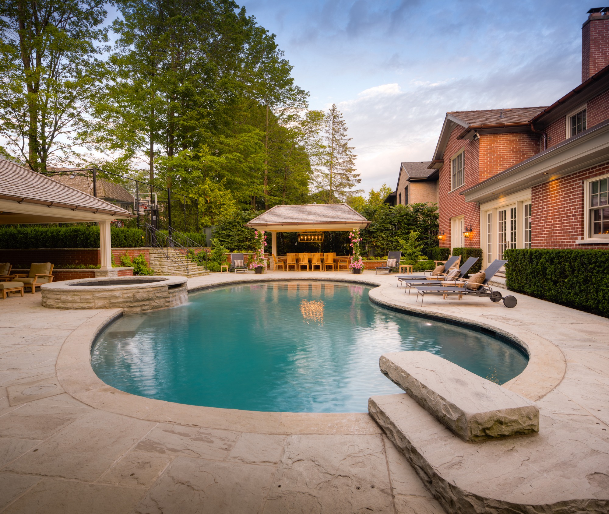 A luxurious backyard with an inground pool, stone patio, outdoor furniture, a covered dining area, and a brick house surrounded by lush trees at dusk.
