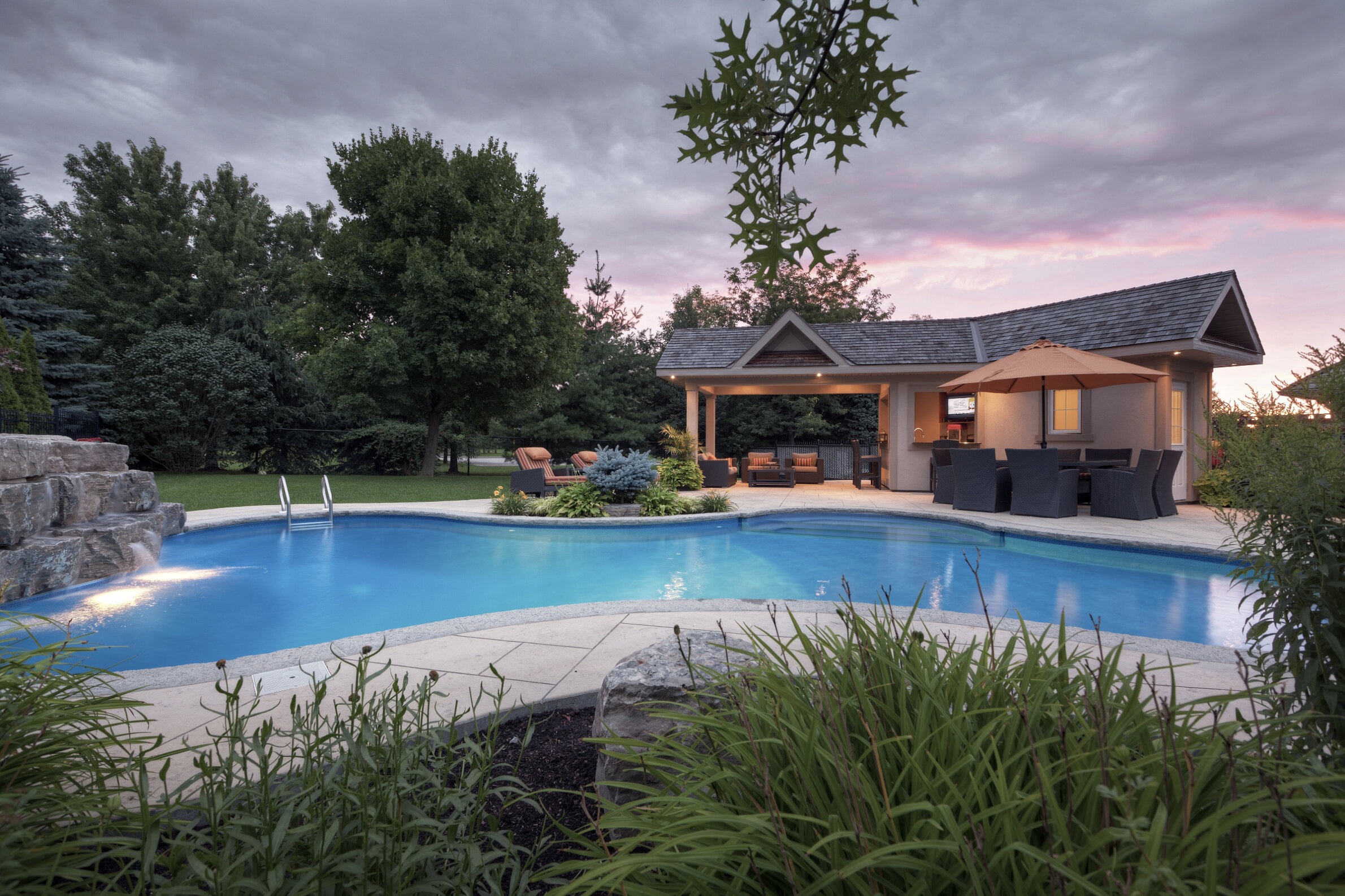 A serene backyard at dusk with an inviting swimming pool, adjacent covered patio with furniture, greenery, and a dramatic twilight sky above.