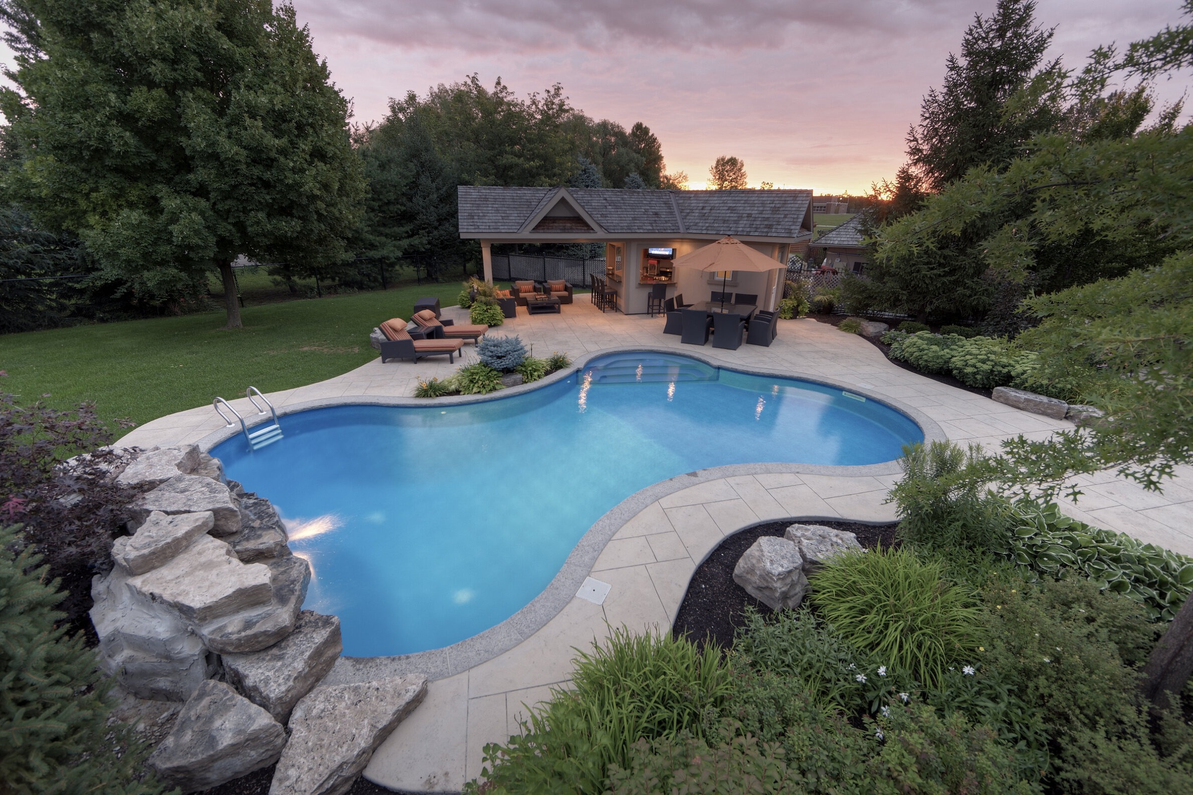 A luxurious backyard with a curved pool at dusk, surrounded by a patio, plush seating, landscaping, and an outdoor kitchen, under a colorful sky.