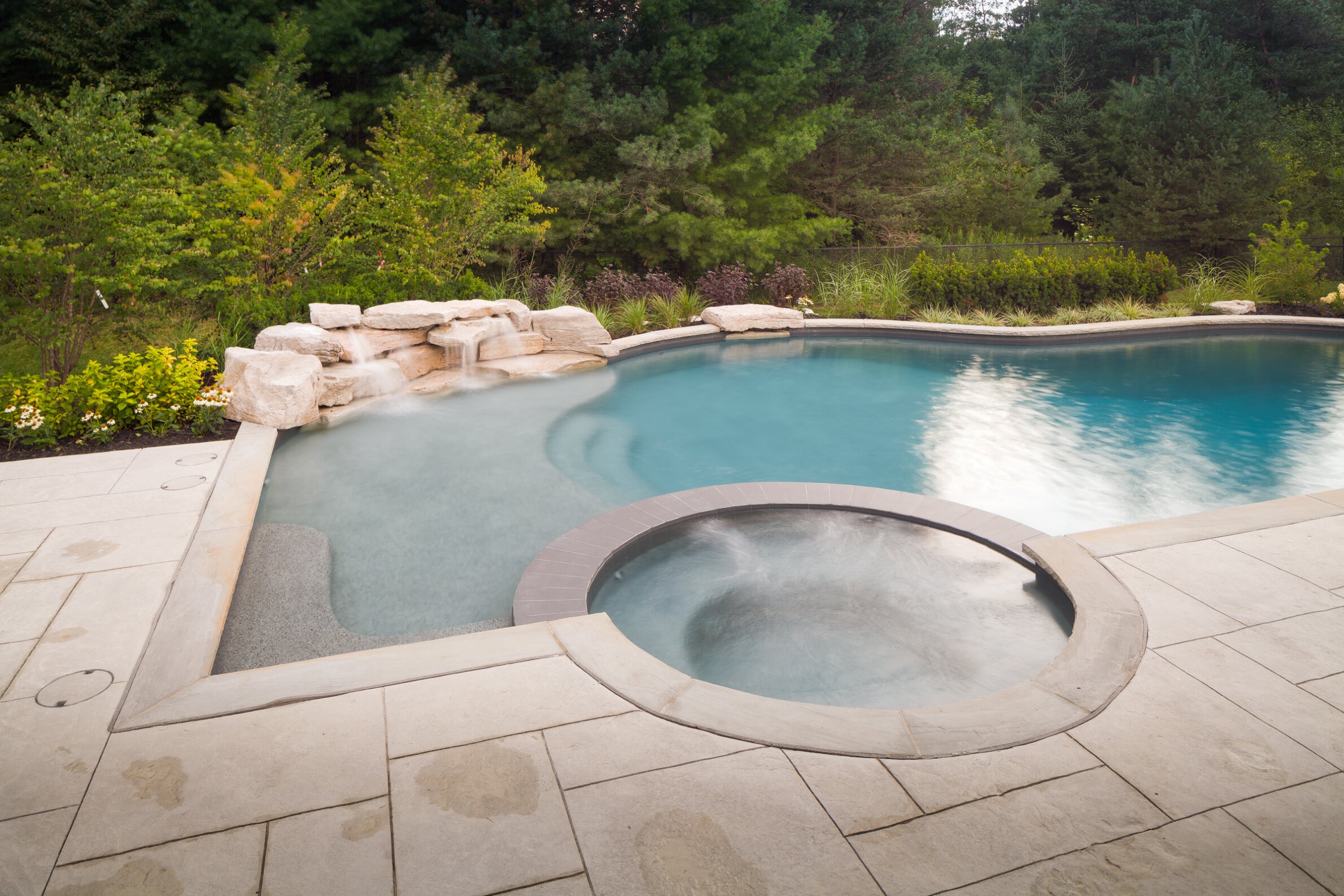 A serene backyard with a curved swimming pool beside a hot tub, featuring a natural rock waterfall, surrounded by lush greenery and a stone deck.