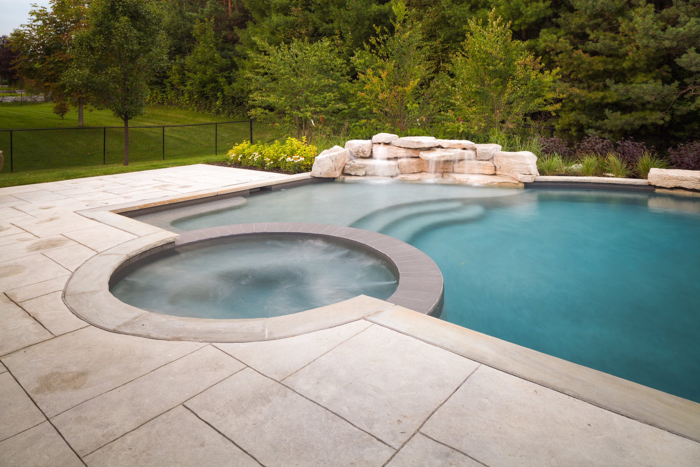 An outdoor swimming pool with an integrated circular hot tub, stone waterfall, surrounded by a patio and landscaping, with a fence in the background.