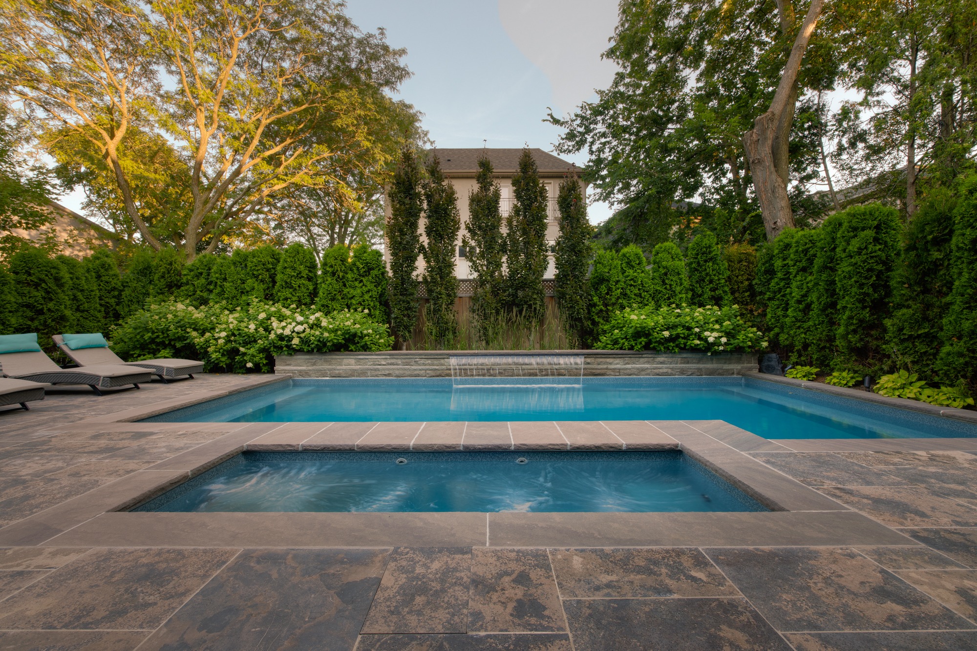 A serene backyard featuring a rectangular swimming pool with an integrated hot tub, surrounded by lush greenery, loungers, and a stone patio at dusk.