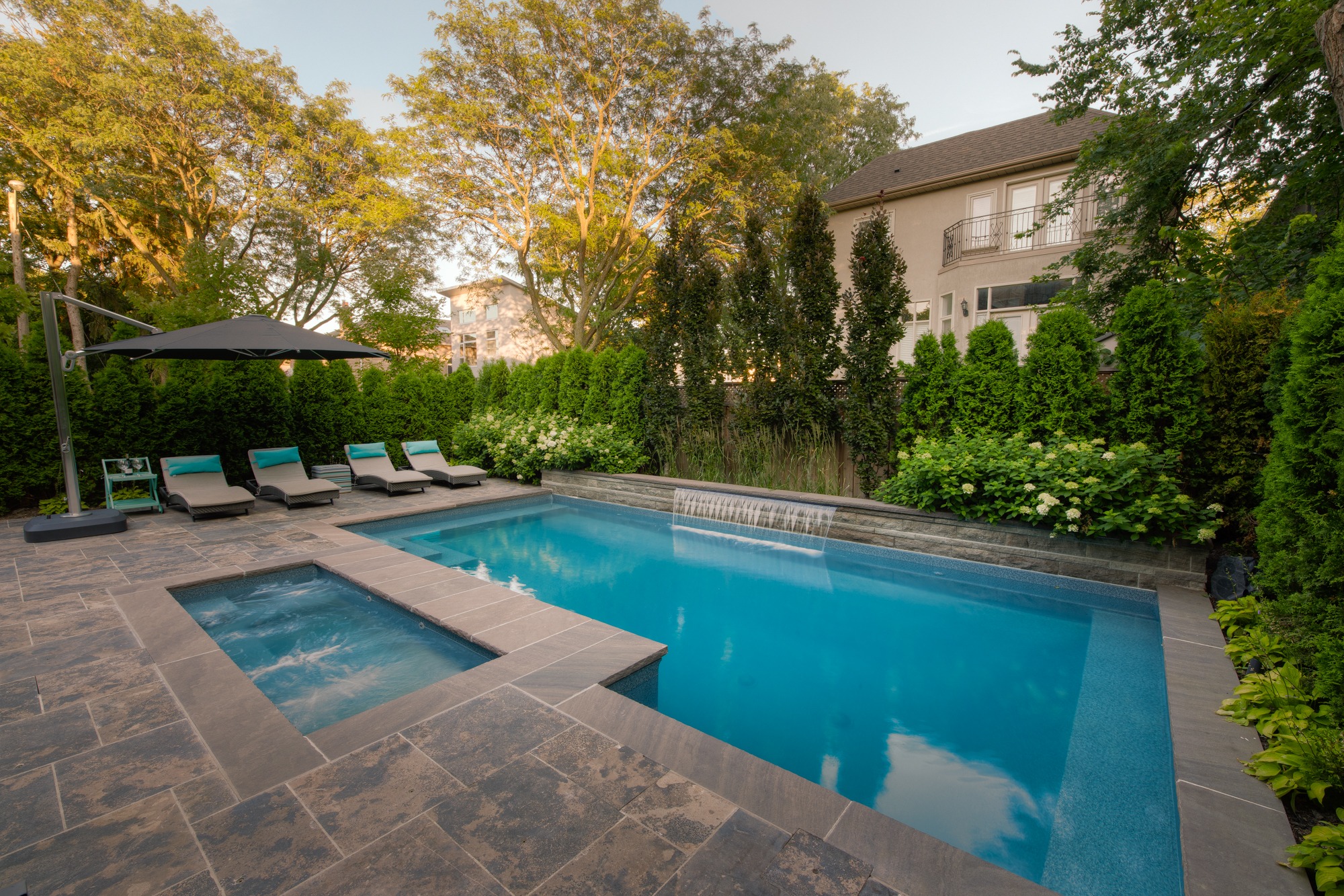 This image shows a luxury backyard with an in-ground swimming pool and jacuzzi, sun loungers under an umbrella, surrounded by a manicured garden and trees.
