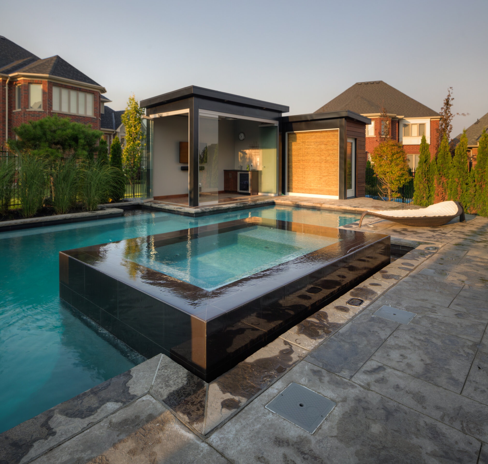 Modern backyard with a luxurious infinity pool and integrated hot tub, surrounded by stone paving, next to a glass-walled pool house at dusk.