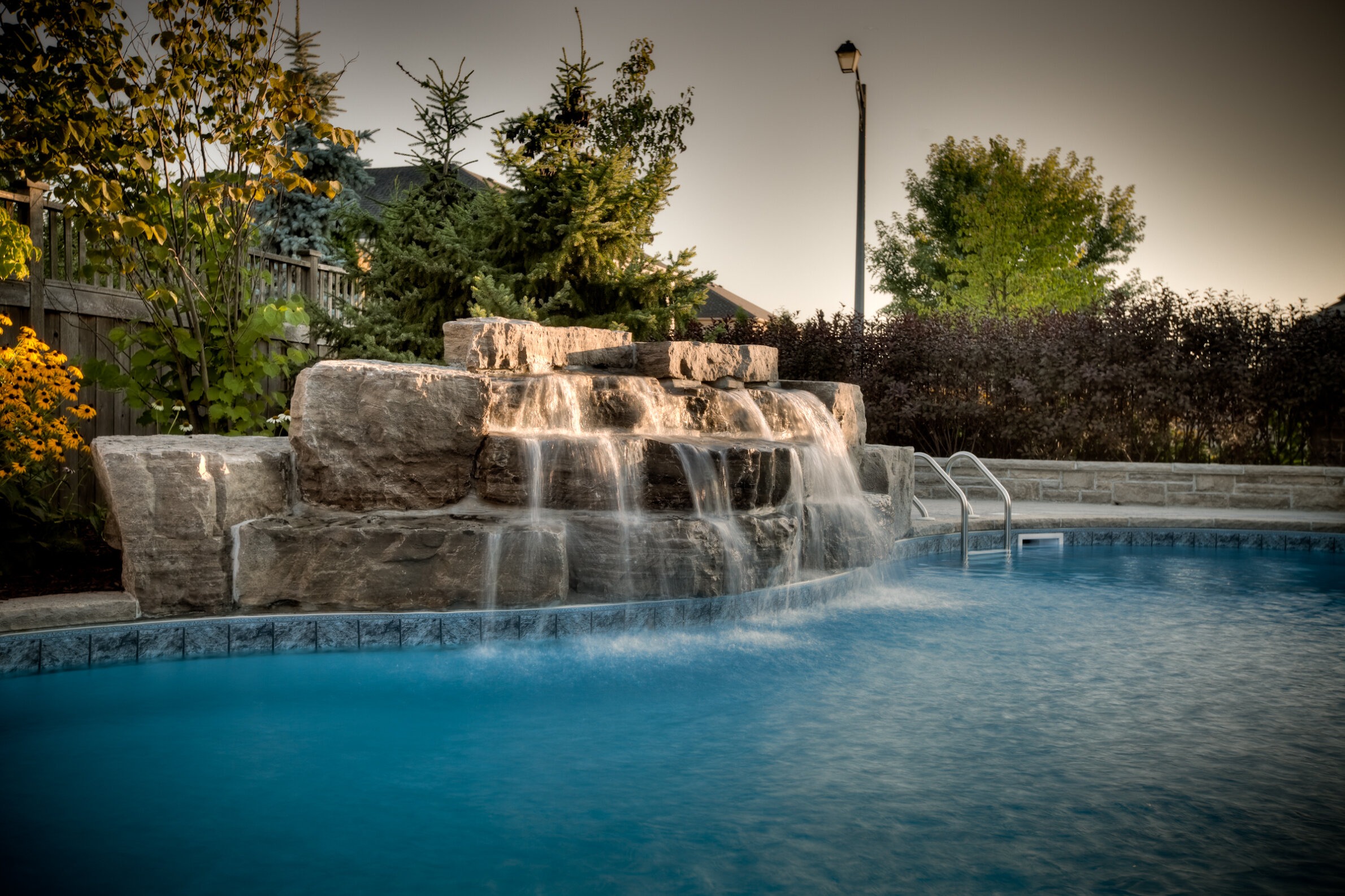 A serene backyard setting with a blue swimming pool featuring a cascading artificial rock waterfall, surrounded by greenery, flowers, and a lamppost.