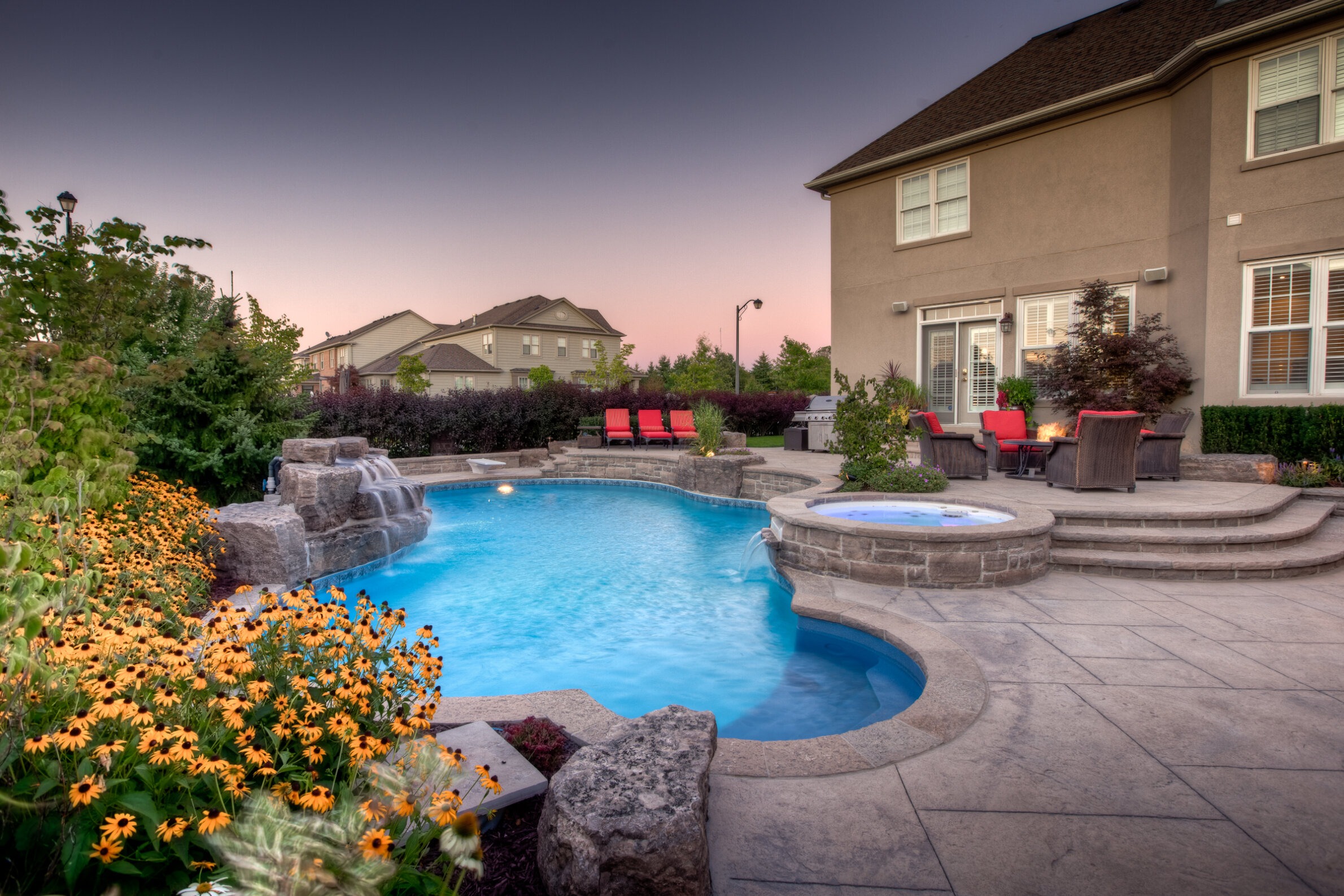 A serene backyard at dusk with an inviting swimming pool, hot tub, waterfall feature, flowering plants, and a cozy seating area with a fire pit.