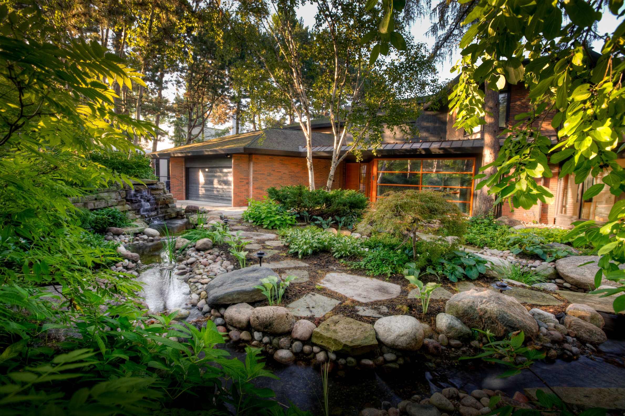A serene residential garden with a waterfall and stream surrounded by lush greenery, leading up to a brick house with large windows nestled among trees.