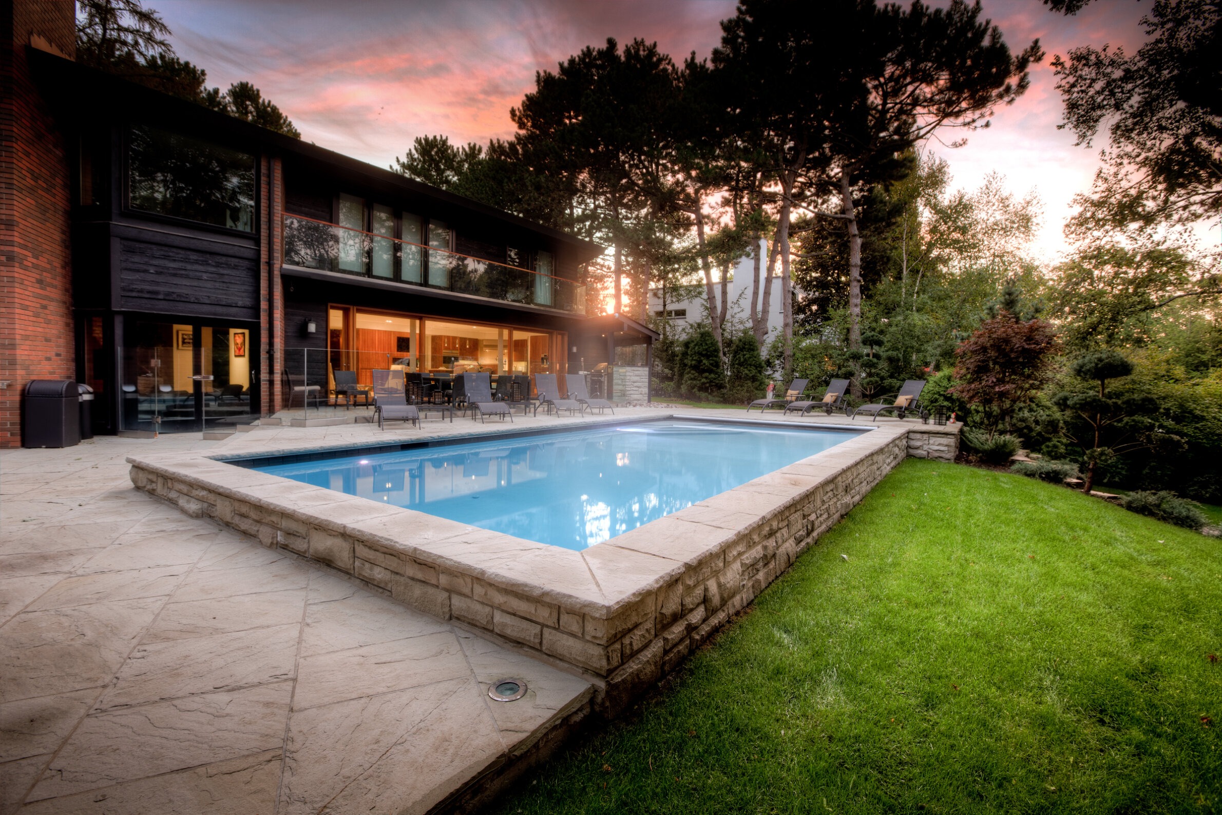 This image shows a luxurious backyard at dusk with a swimming pool, lounge chairs, and a modern house with large windows overlooking a landscaped garden.