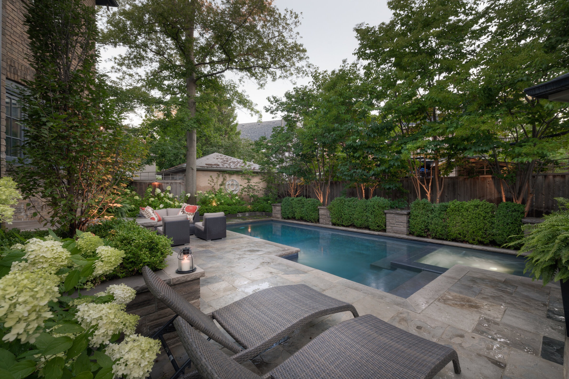 An inviting backyard with a pool, stone patio, outdoor furniture, lush greenery, and a lantern, exuding a serene, relaxing ambiance in a residential setting.