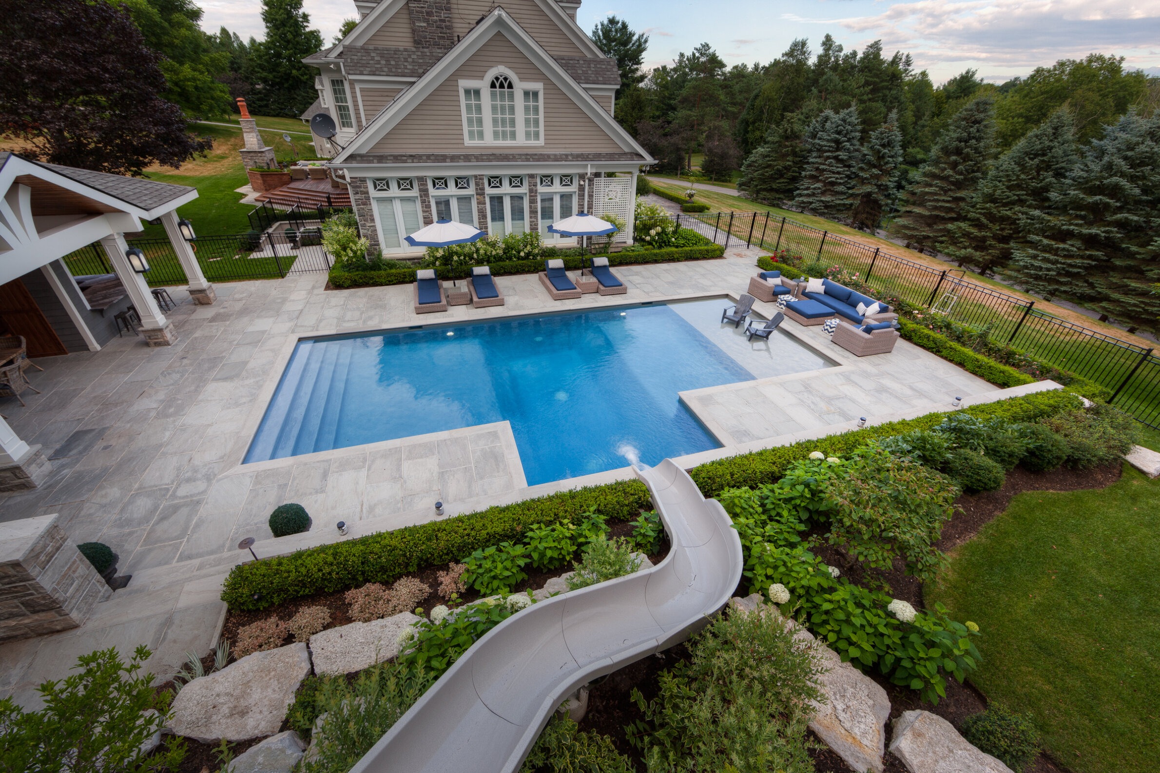 This image shows a luxurious backyard with a swimming pool, lounge chairs, a slide, landscaping, a gazebo, and a large house, all enclosed by a fence.