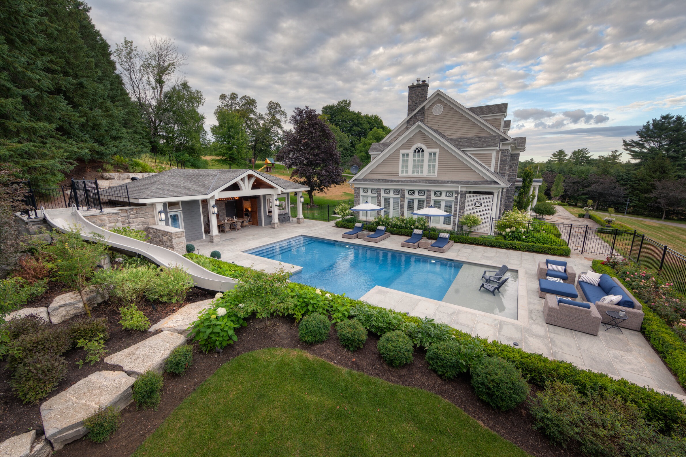 An upscale backyard with a swimming pool, lounge chairs, slide, manicured gardens, pool house, and a spacious two-story home against a cloudy sky.