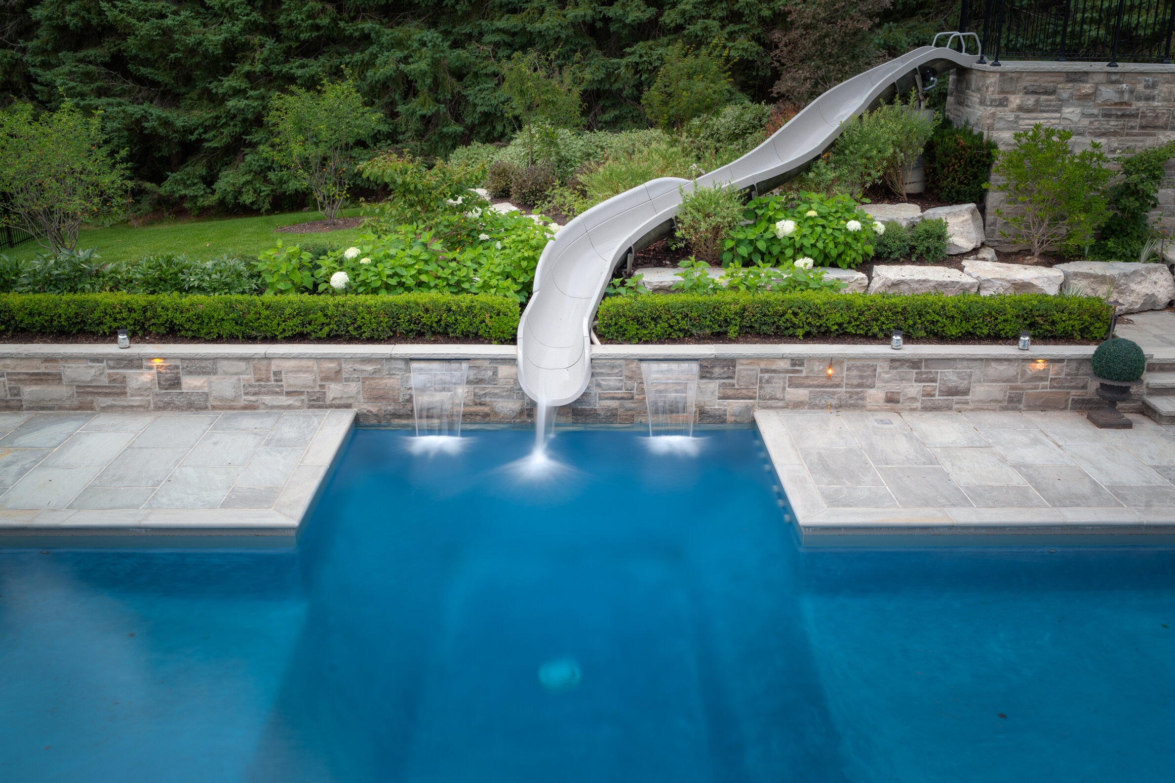 A residential outdoor pool with a curved slide overlooking lush greenery and foliage, featuring stone landscaping and subtle outdoor lighting.