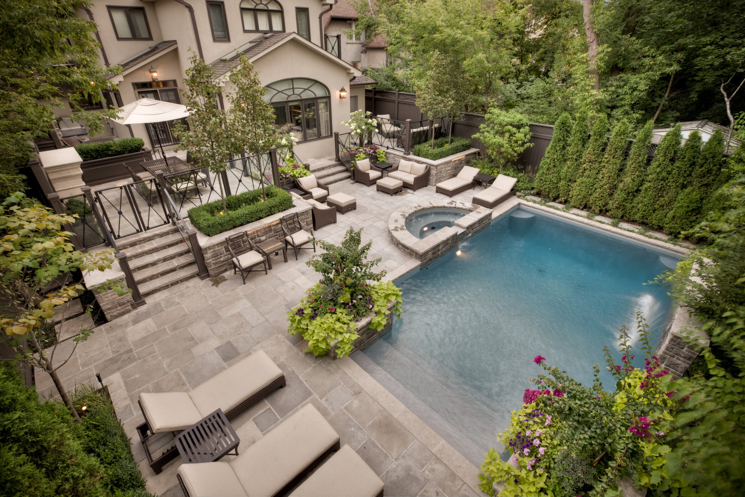 An upscale backyard with a swimming pool, hot tub, outdoor dining area, lounge chairs, stone patio, lush greenery, and a beige stucco house.