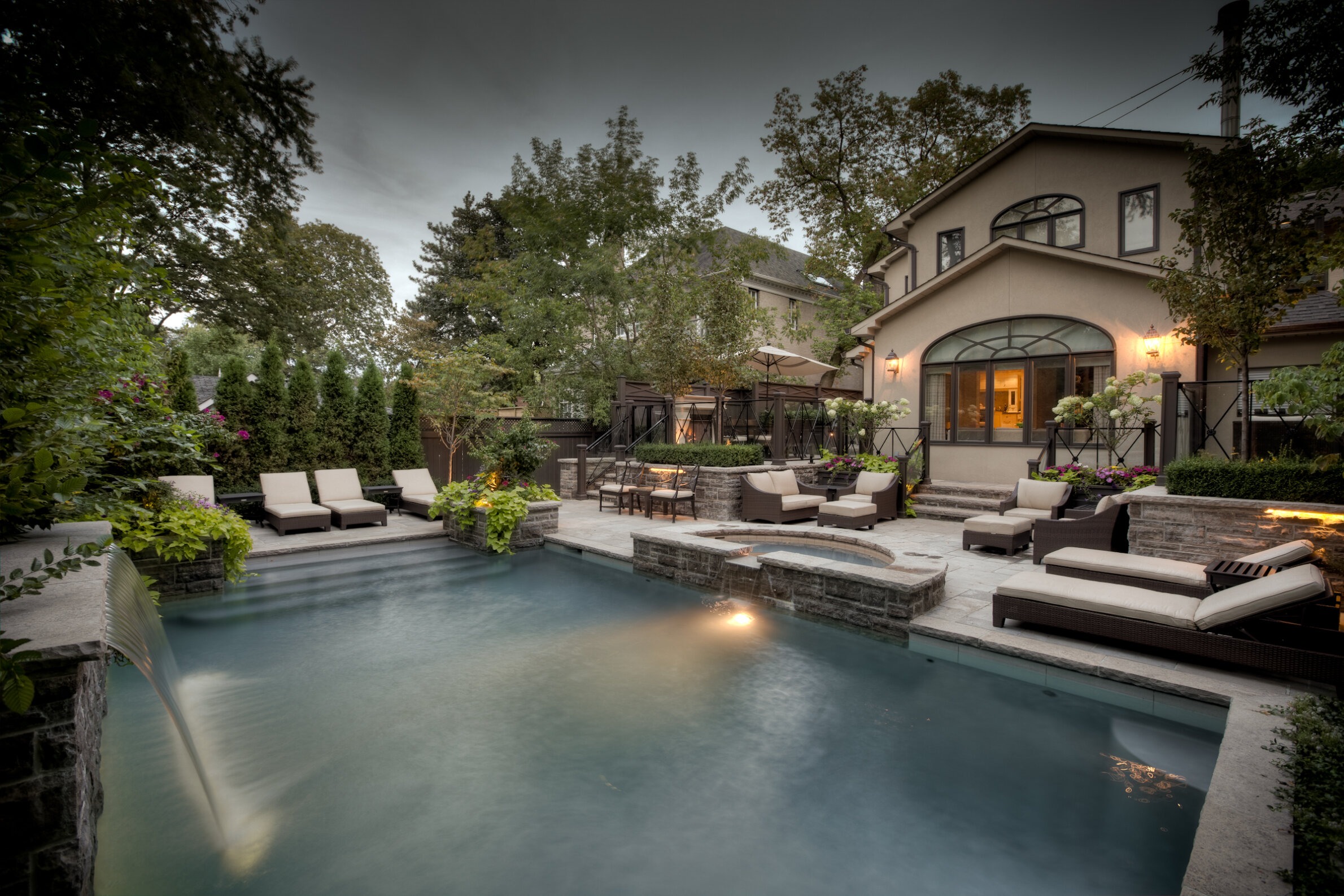 An upscale backyard with a swimming pool, elegant outdoor furniture, lush landscaping, stonework, and a warmly lit residence in a serene twilight setting.