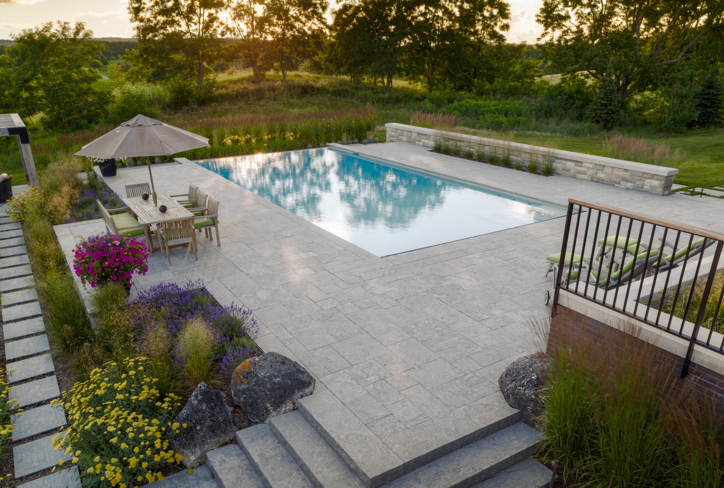 This is an image of a serene backyard with a rectangular swimming pool, outdoor furniture under an umbrella, stone paving, and colorful landscaping.