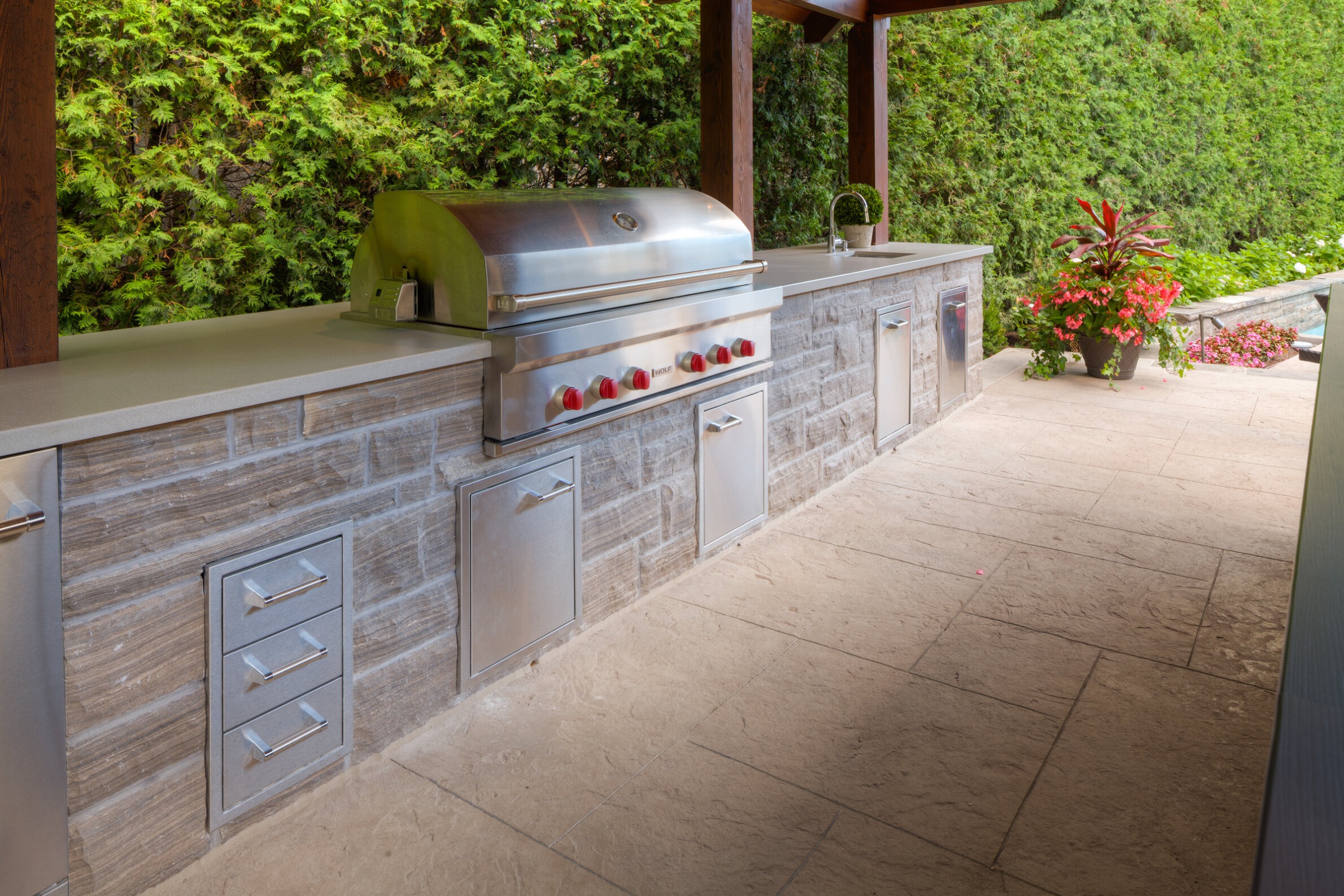 An outdoor kitchen with a stainless steel barbecue grill, storage drawers, a sink, and a counter set against a lush green backdrop and tiled floor.