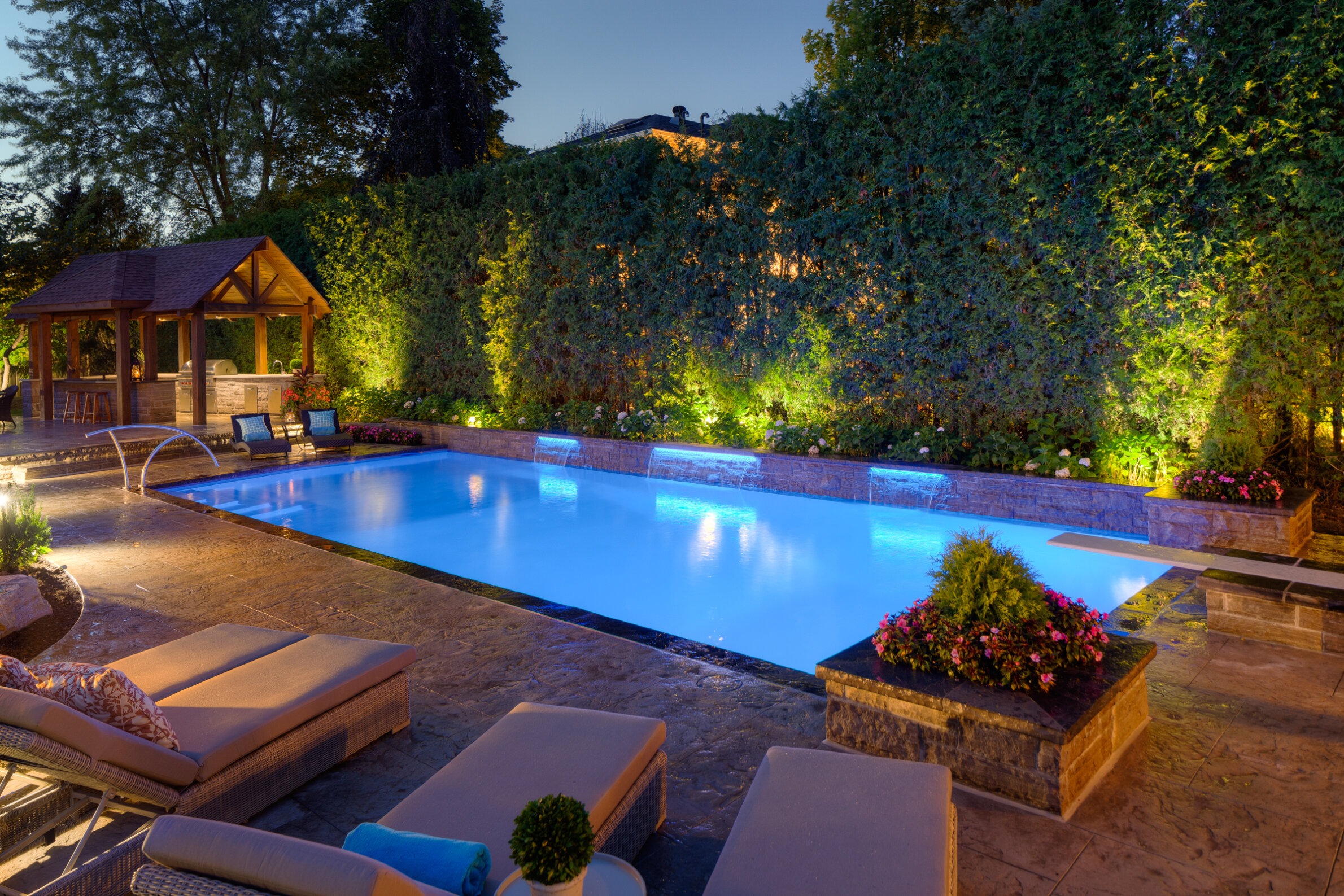 An outdoor swimming pool illuminated at dusk with underwater lights, surrounded by loungers, plants, a gazebo, and a stone patio.