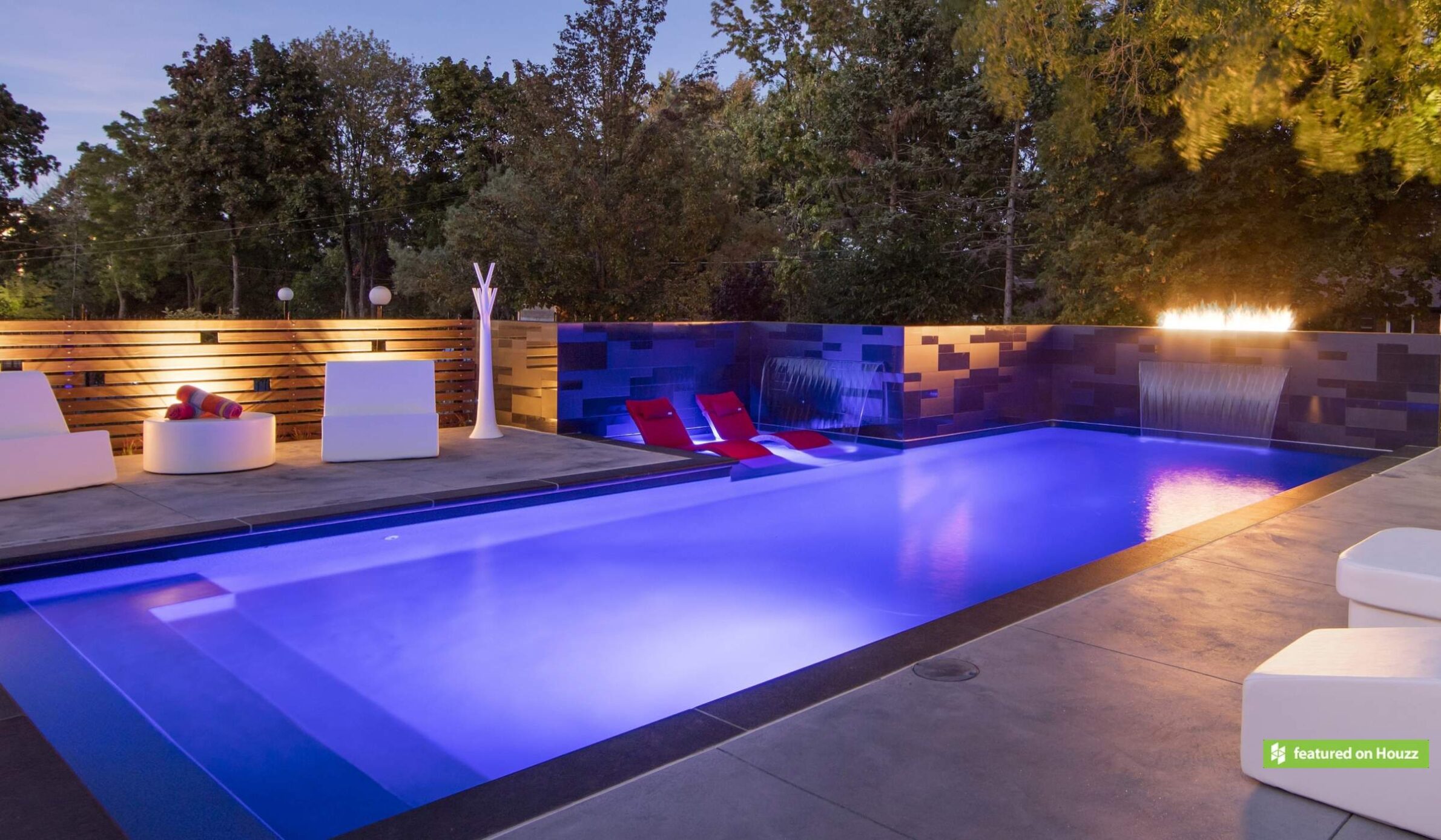 A modern outdoor swimming pool with blue lighting at dusk, featuring loungers, a waterfall, and a stylish seating area with wooden fencing.
